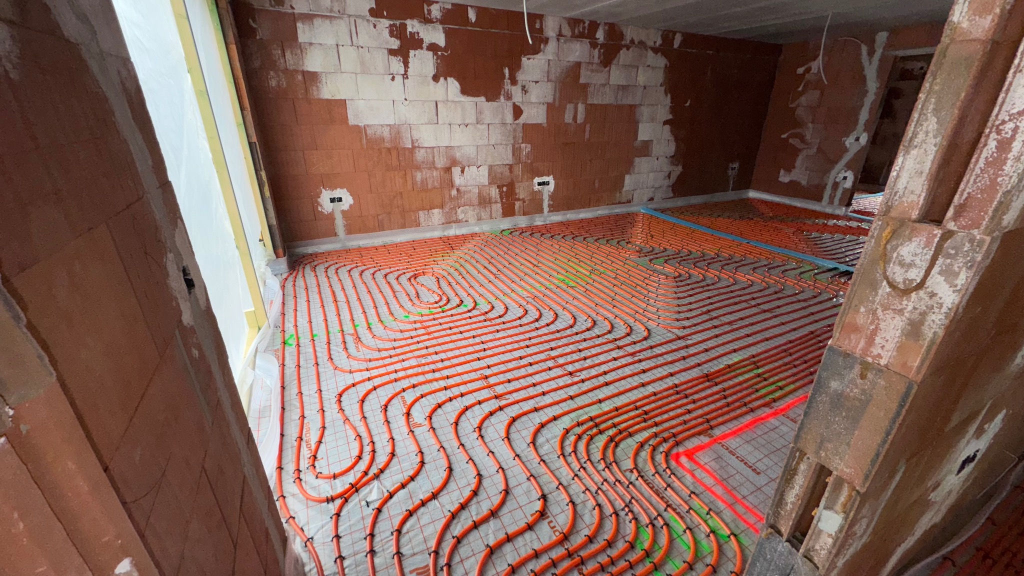 Pomieszczenie w trakcie montażu ogrzewania podłogowego z rurkami ułożonymi na folii z siatką. Ściany z cegły, widoczne instalacje elektryczne.