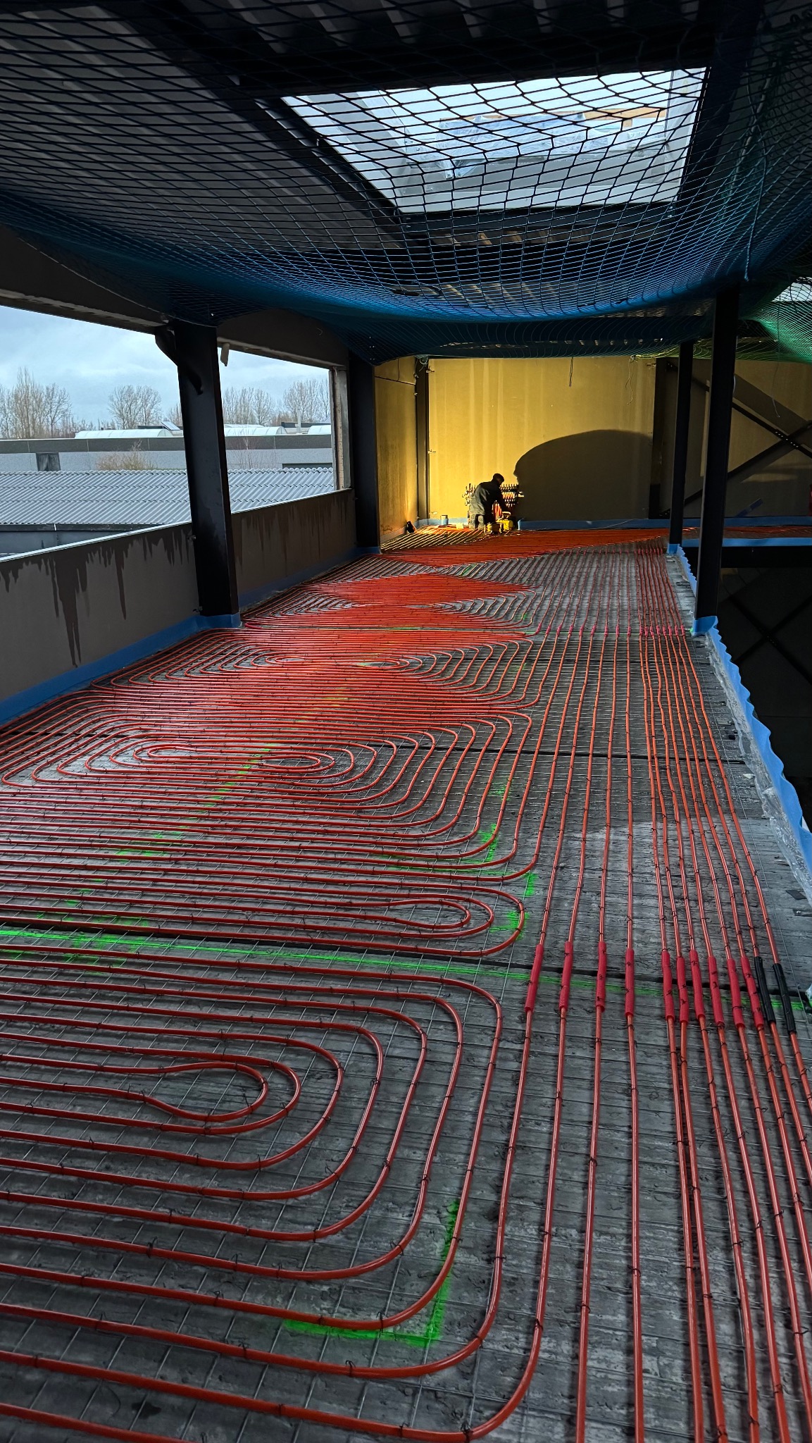 Instalacja ogrzewania podłogowego z czerwonymi rurami na metalowej siatce, widok z góry, w tle osoba przyłącza rury w budynku w stanie surowym.