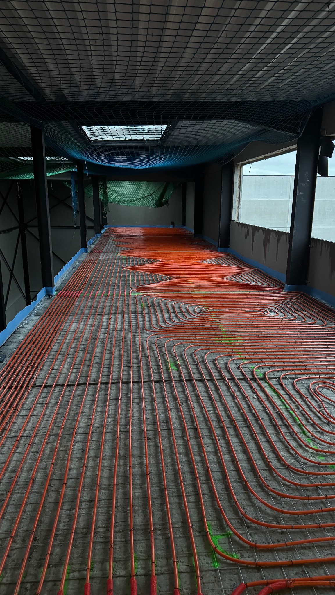 Pomarańczowe rury ogrzewania podłogowego ułożone na szarym betonie w budynku w stanie surowym, widoczne okno i siatka zabezpieczająca.