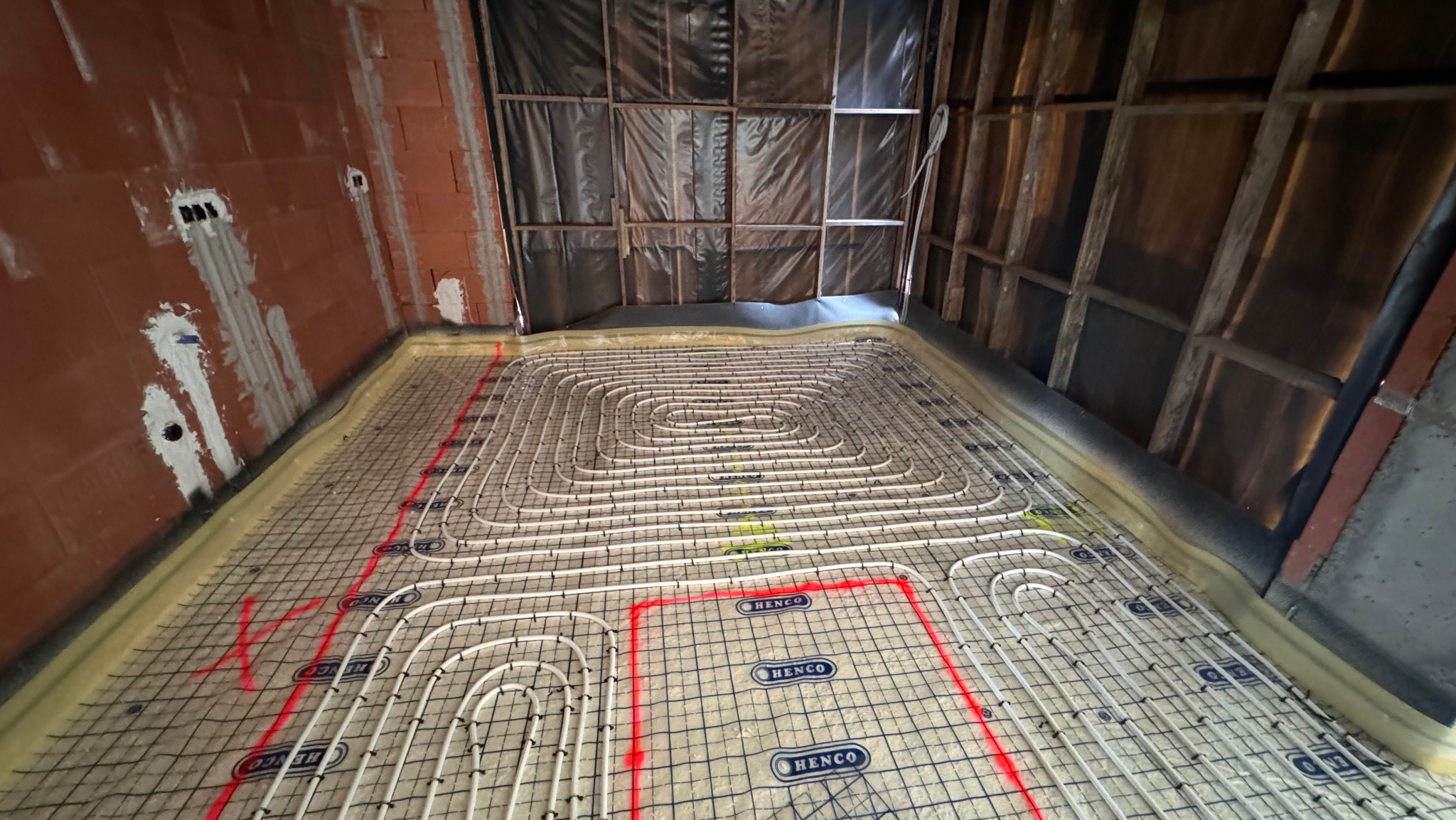 Instalacja ogrzewania podłogowego Henco na izolacji, widoczne rury w kształcie spirali i czerwone oznaczenia. Surowy stan ścian i stropu.
