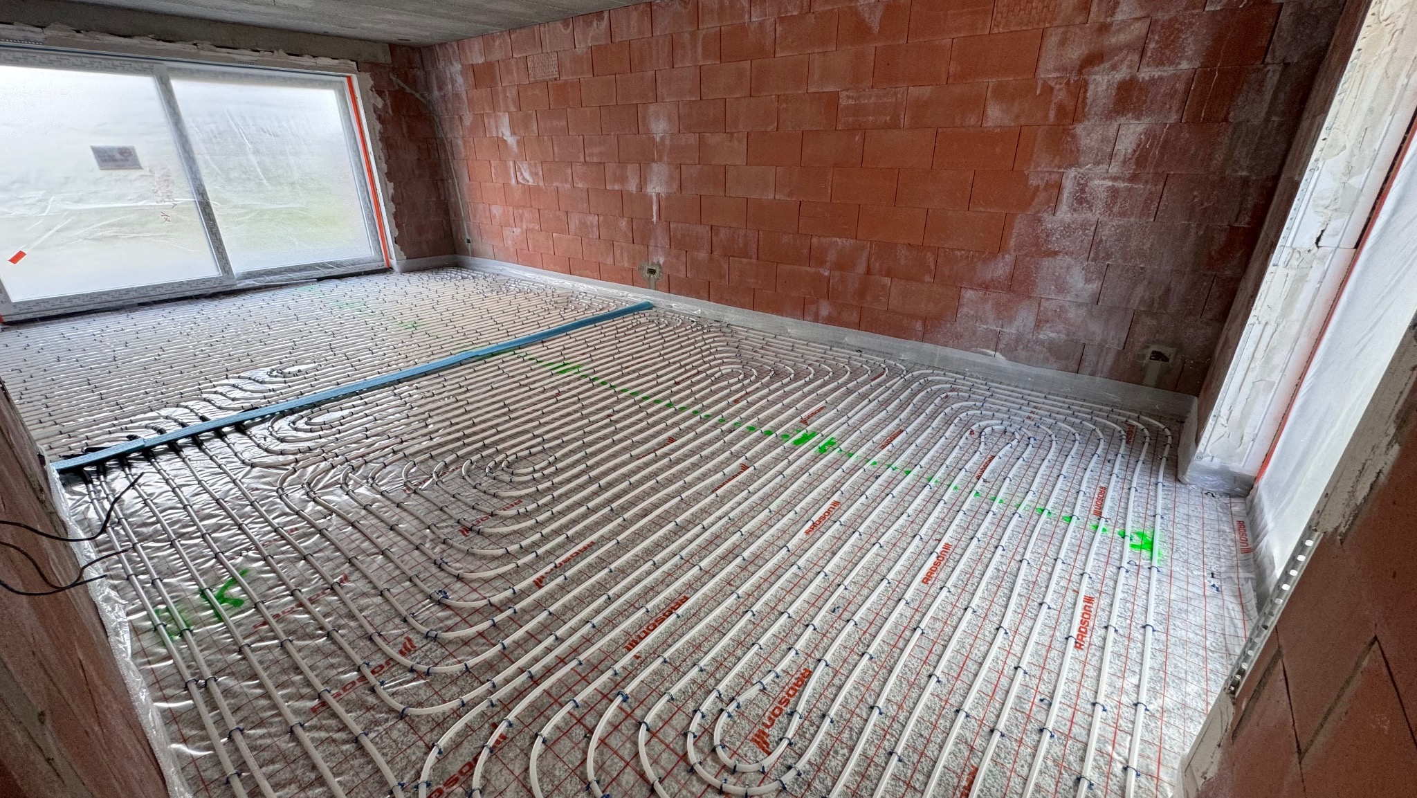 Białe rury ogrzewania podłogowego ułożone spiralnie na folii w surowym pomieszczeniu z ceglanymi ścianami i oknem, przed wylaniem posadzki. Widoczne zielone oznaczenia.