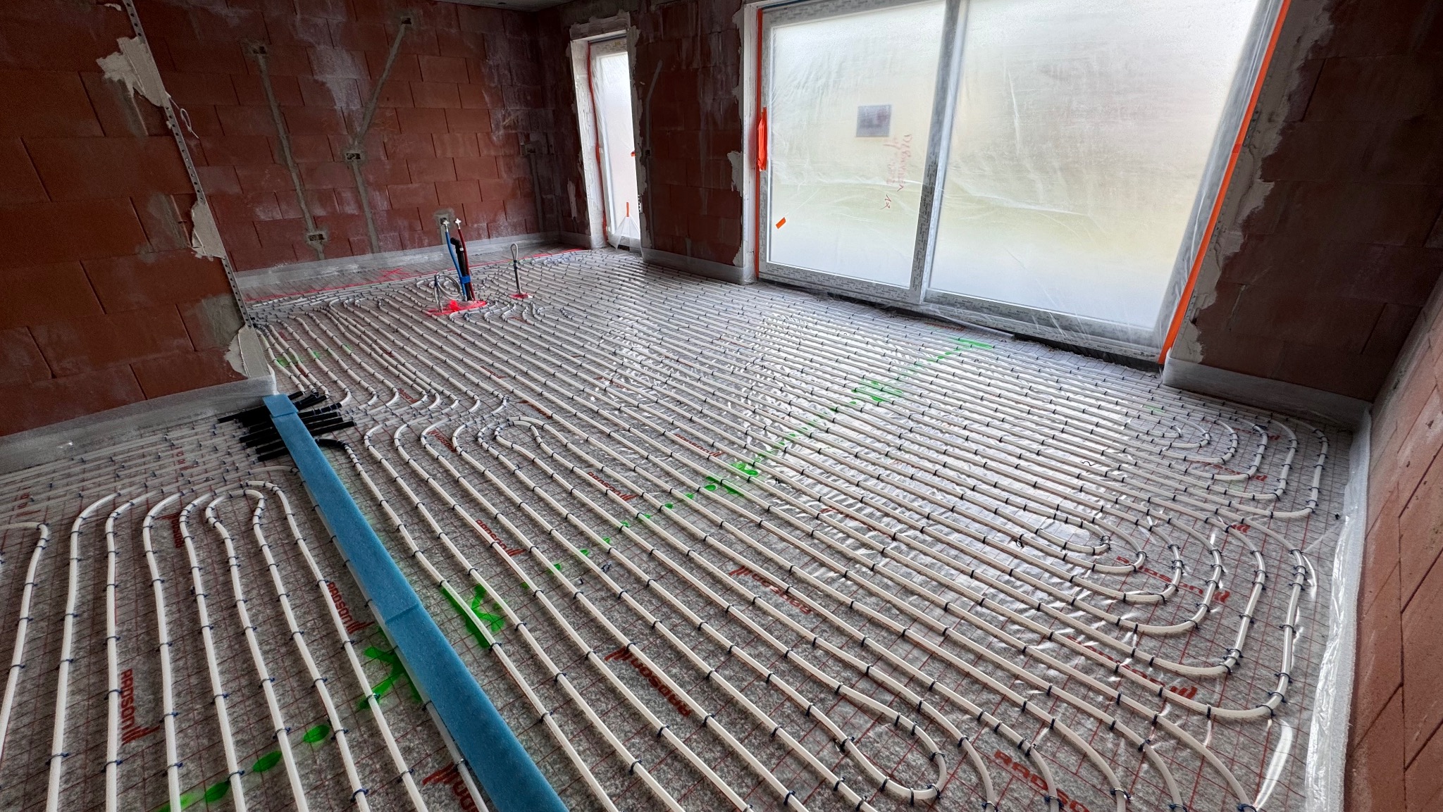 Ułożone rury ogrzewania podłogowego na folii izolacyjnej w surowym wnętrzu z ceglanymi ścianami i oknami zabezpieczonymi folią. Widoczny grzebień do mocowania rur.
