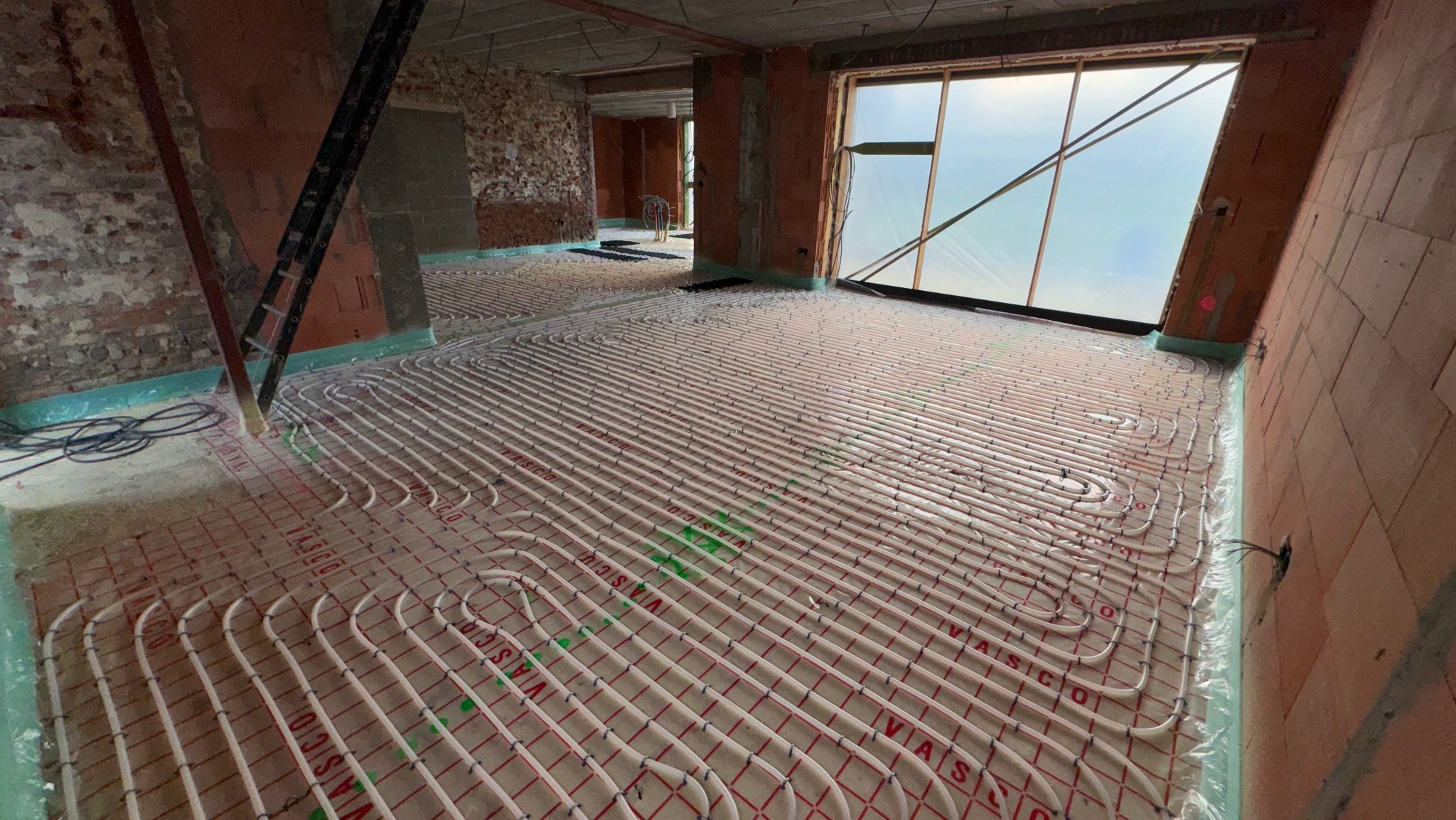 Białe rury ogrzewania podłogowego ułożone na zielonej folii w surowym wnętrzu, z widocznym oknem i fragmentem drabiny, gotowe do zalania posadzką.