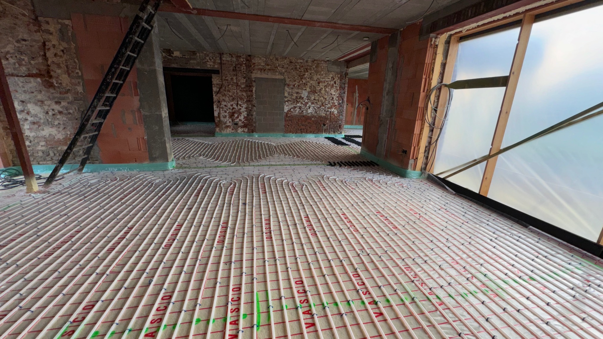 Instalacja ogrzewania podłogowego w surowym wnętrzu z ceglanymi ścianami. Równoległe rury na izolacji, drabina oparta o ścianę, widok na okno.