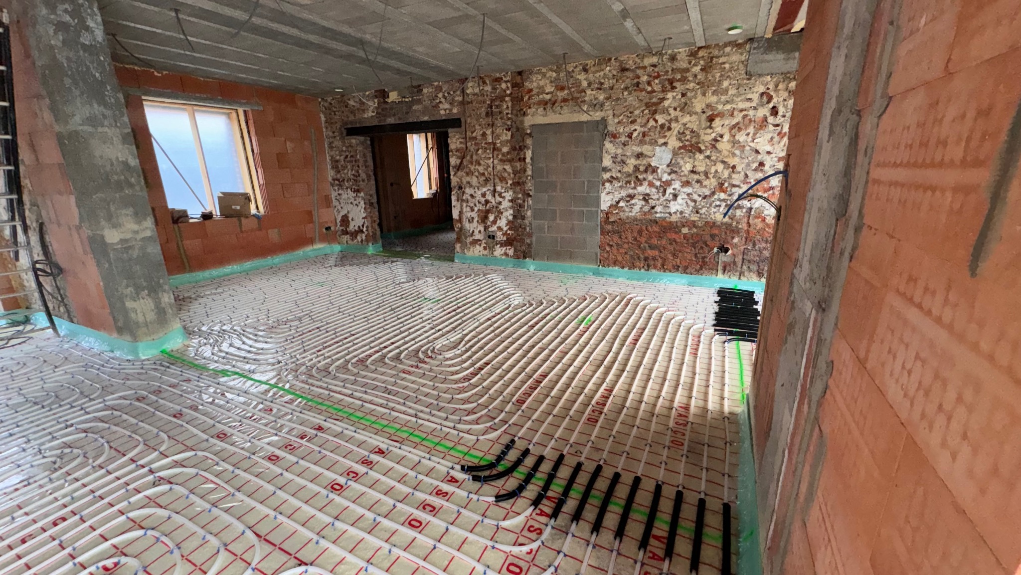 Białe rury ogrzewania podłogowego ułożone na izolacji w surowym wnętrzu z ceglanymi ścianami i oknem. Widoczne czarne złączki i zielona folia izolacyjna.