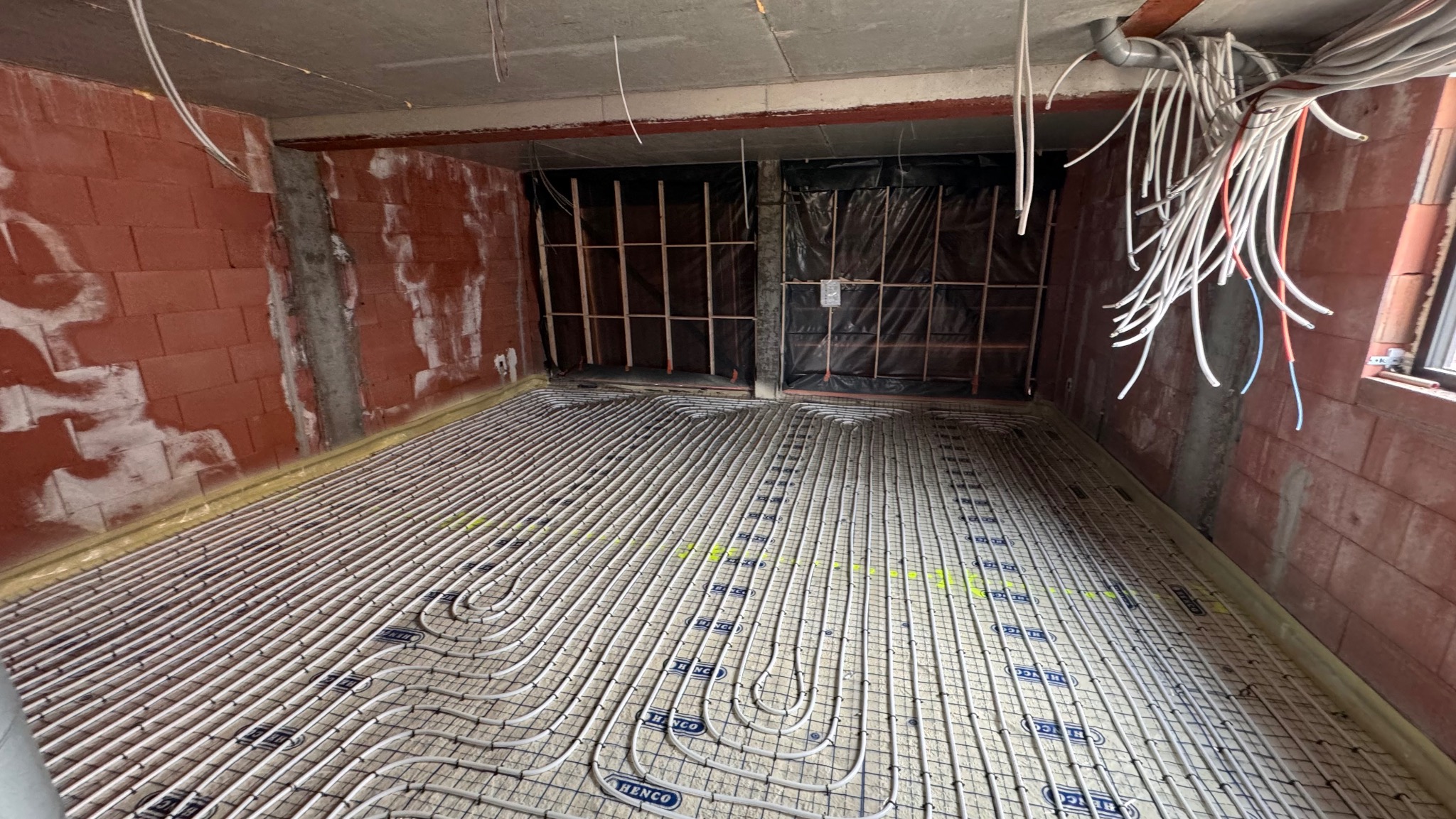 Instalacja ogrzewania podłogowego Henco na izolacji, widoczne rury i przewody elektryczne. Surowy stan, ściany z czerwonej cegły.