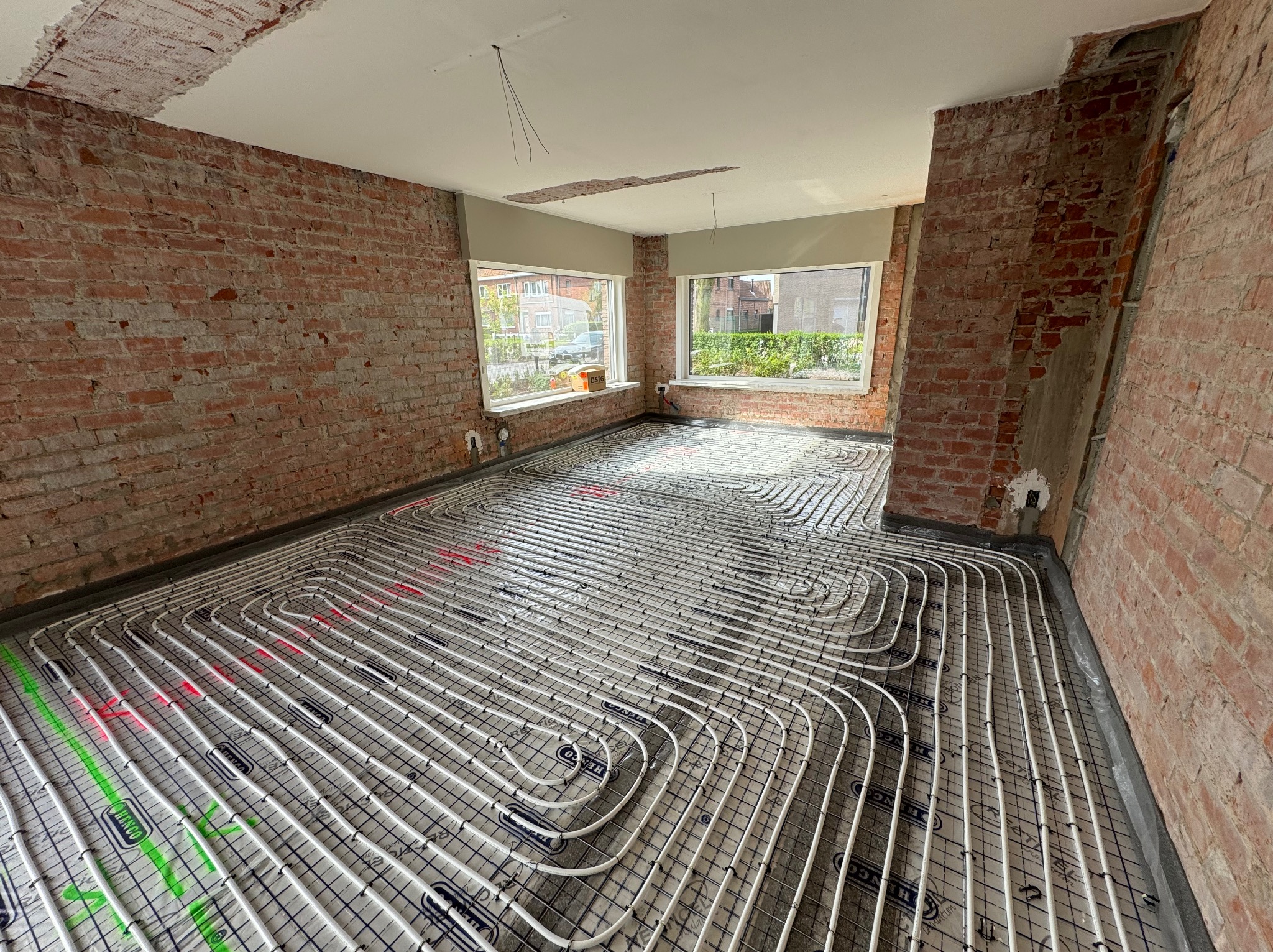 Rozłożona instalacja ogrzewania podłogowego na folii z siatką, w surowym wnętrzu z ceglanymi ścianami, widok na okno i ulicę.