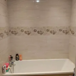 Wykończona łazienka z beżowymi płytkami i dekoracyjnym pasem z motywem liści. Wanna w zabudowie. Kosmetyki na wannie.