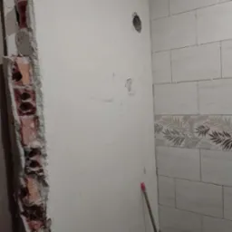 Fragment ściany z odsłoniętą cegłą i częściowo położonymi, jasnymi płytkami z motywem roślinnym. Widoczne krzyżyki dystansowe. Etap prac wykończeniowych.