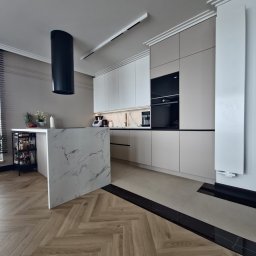 R Design Meble Na Wymiar - Nowoczesna kuchnia z wyspą wykończoną marmurem, fronty szafek w odcieniach bieli i beżu, czarny okap cylindryczny, parkiet w jodełkę i czarne akcenty na podłodze.