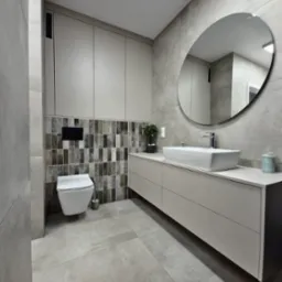 Nowoczesna łazienka w szarościach z wiszącą toaletą, umywalką nablatową i okrągłym lustrem. Ciekawy akcent stanowią płytki dekoracyjne za toaletą.