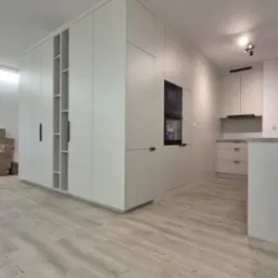 Jasna, nowoczesna kuchnia z zabudową na wymiar, łącząca szafy i część kuchenną z piekarnikiem. Minimalistyczne uchwyty, drewnopodobna podłoga, neutralna kolorystyka.