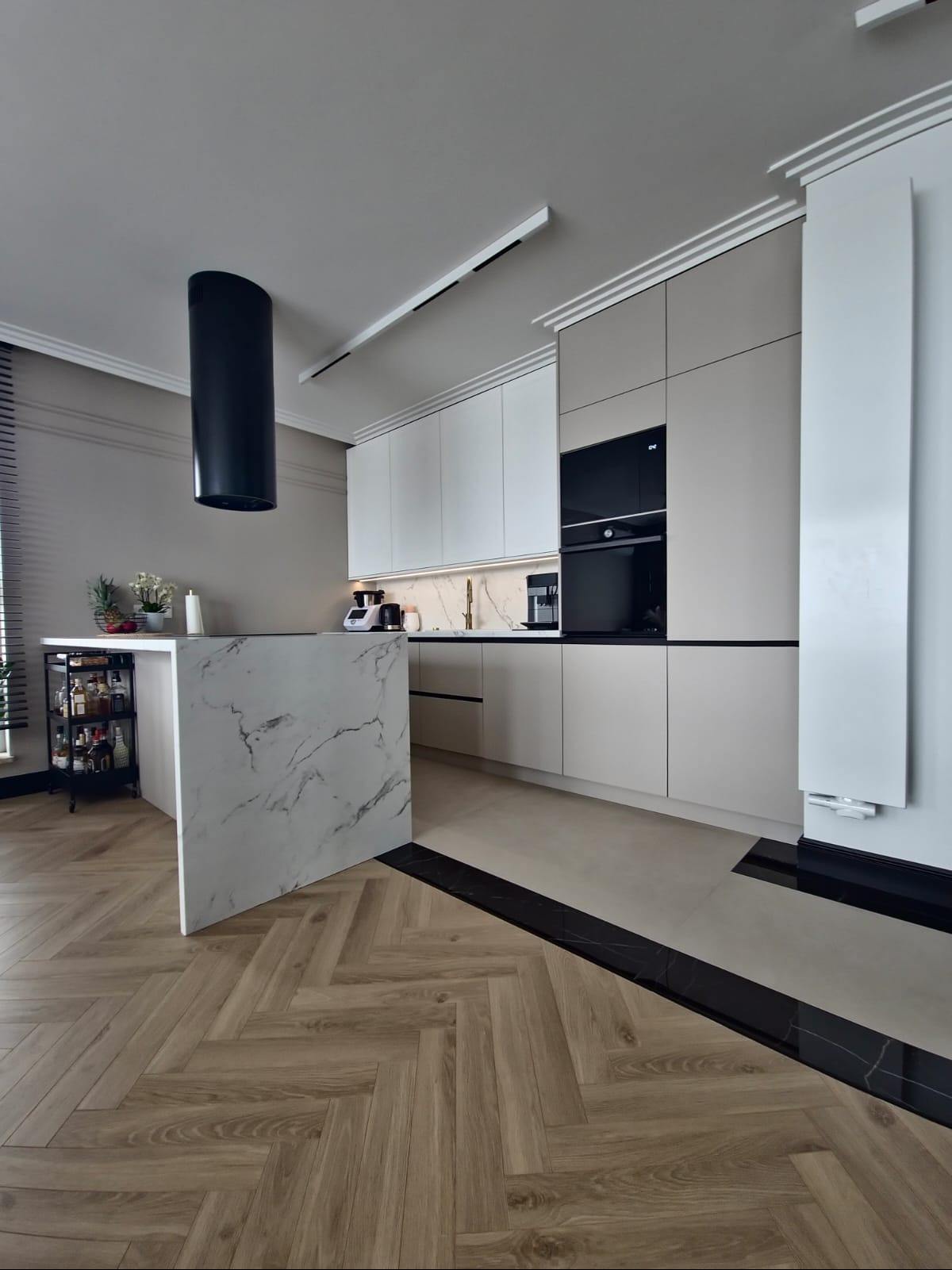 Nowoczesna kuchnia z wyspą wykończoną marmurem, fronty szafek w odcieniach bieli i beżu, czarny okap cylindryczny, parkiet w jodełkę i czarne akcenty na podłodze.