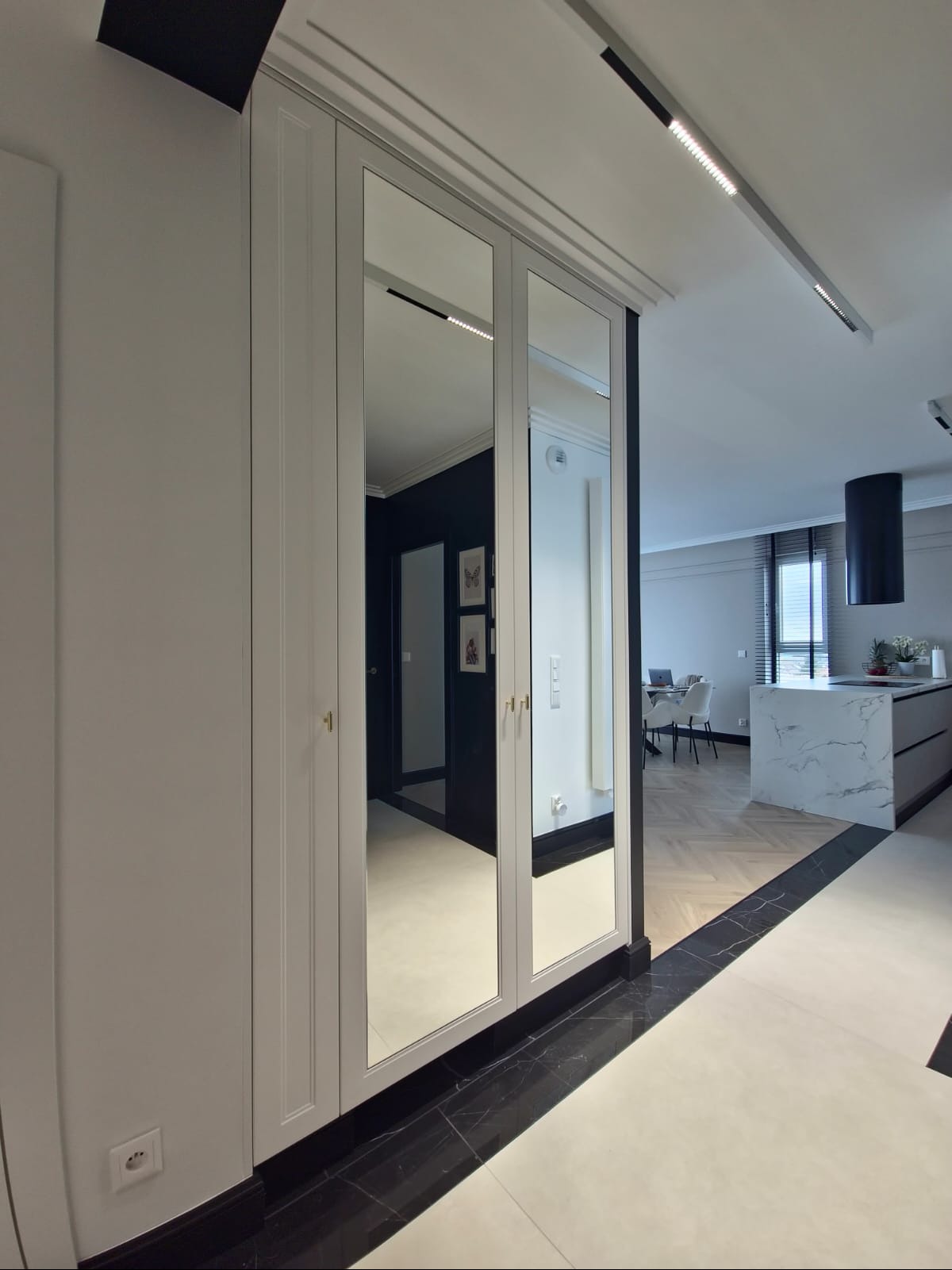 Elegancka szafa wnękowa z lustrzanymi drzwiami odbija wnętrze nowoczesnej kuchni z wyspą i parkietem w jodełkę, tworząc wrażenie przestronności.