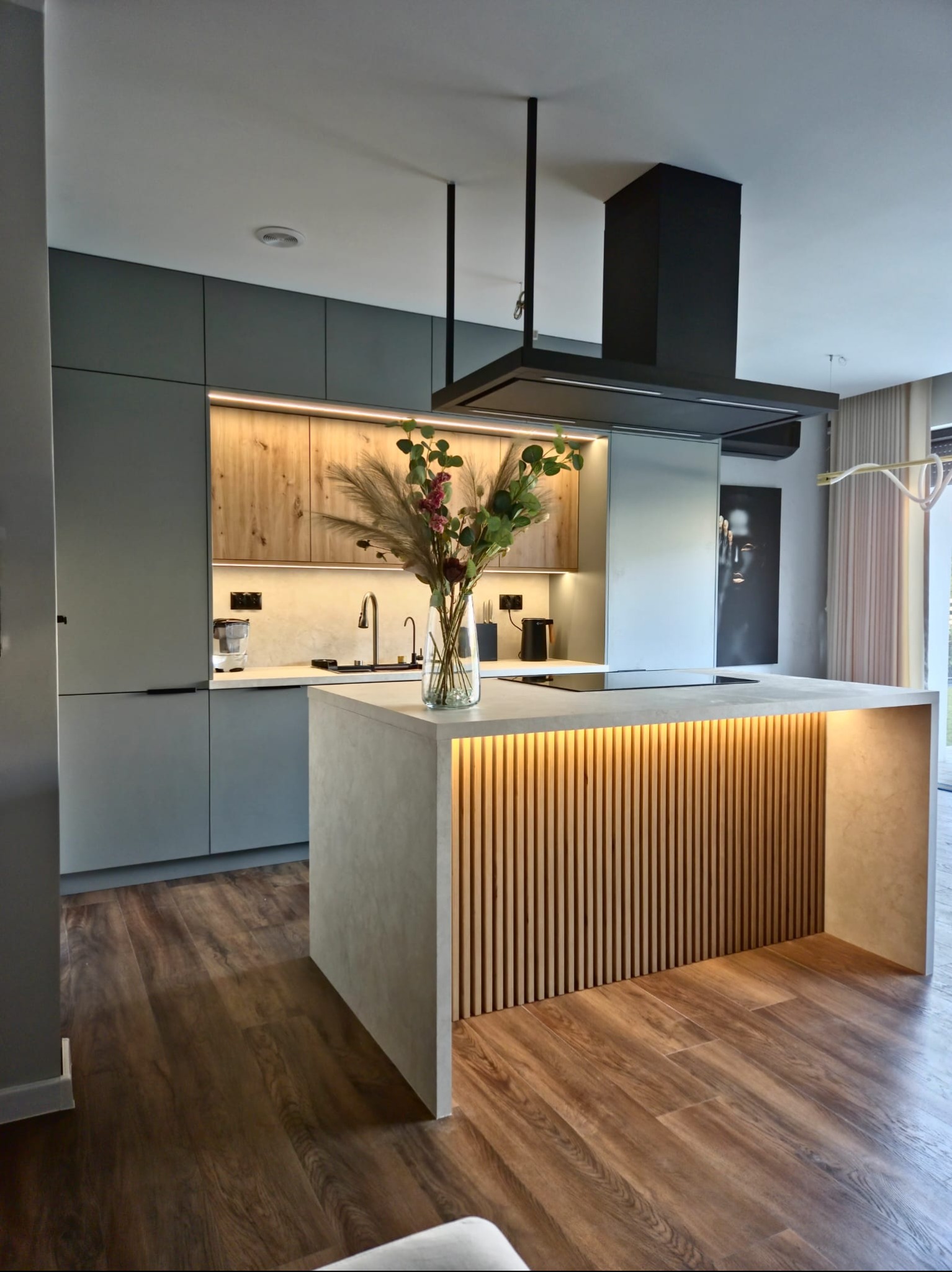 Nowoczesna kuchnia z wyspą oświetloną listwami LED, szare szafki i drewniane akcenty, minimalistyczny okap, drewniana podłoga i wazon z kwiatami na blacie.