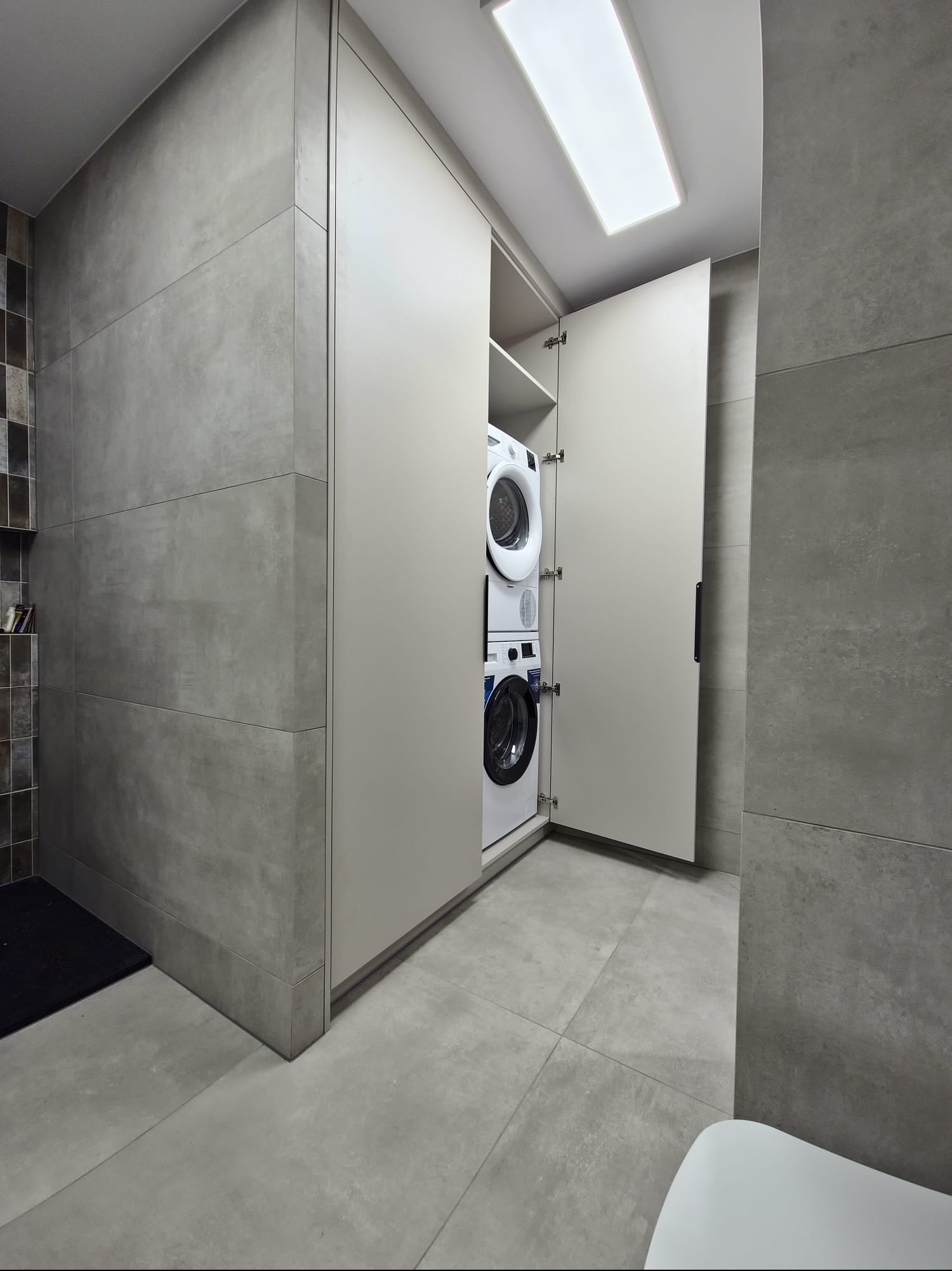 Szara łazienka z szafą na wymiar, w której umieszczono pralkę i suszarkę. Otwarta szafa z beżowymi frontami, szare płytki na ścianach i podłodze.
