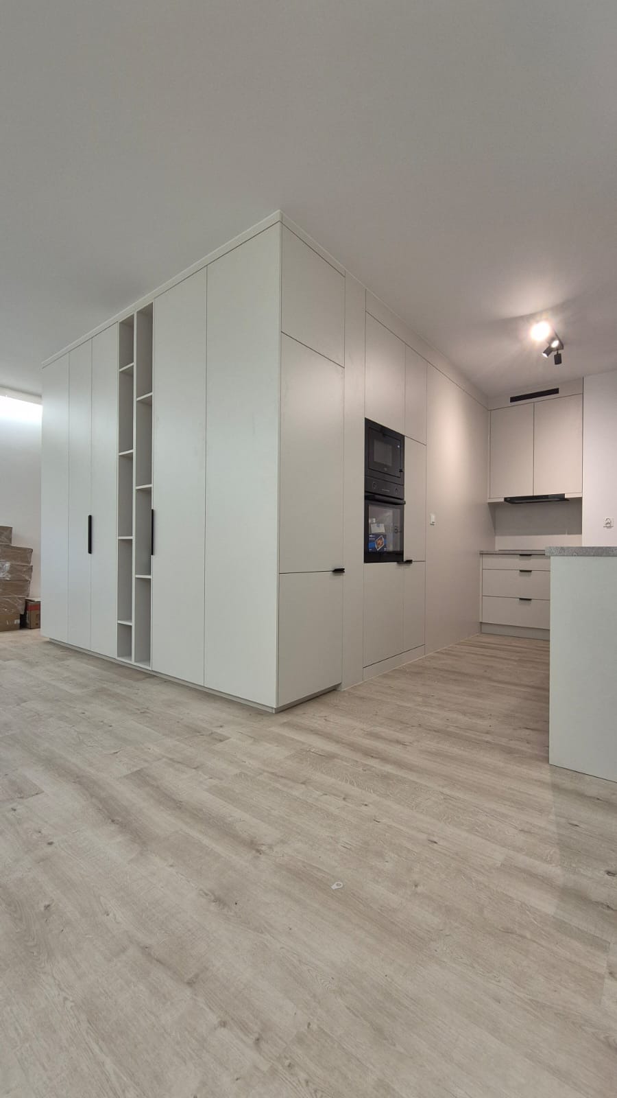 Jasna, nowoczesna kuchnia z zabudową na wymiar, łącząca szafy i część kuchenną z piekarnikiem. Minimalistyczne uchwyty, drewnopodobna podłoga, neutralna kolorystyka.