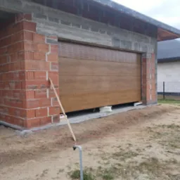 Montaż brązowej bramy garażowej segmentowej w nowym budynku z czerwonej cegły. Brama częściowo otwarta, widoczne elementy montażowe i materiały izolacyjne.