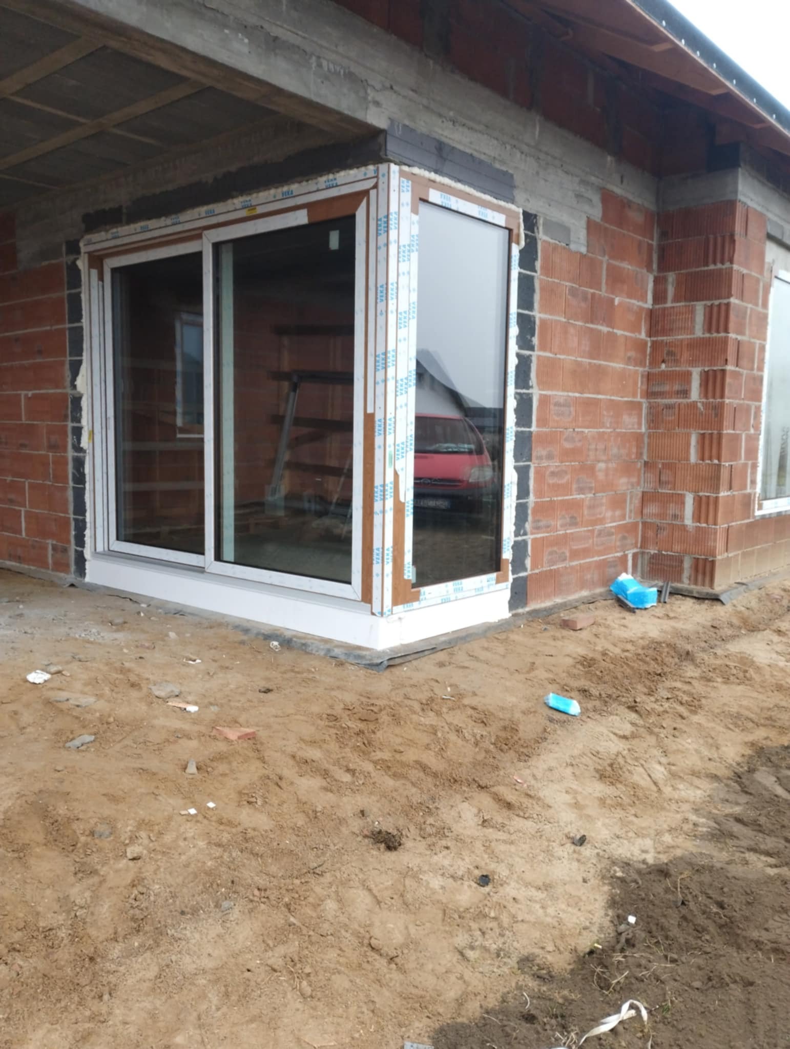 Narożne okno PCV w surowym murze z czerwonej cegły, zabezpieczone folią. Widoczne ślady pianki montażowej i gruzu. W tle czerwony samochód.