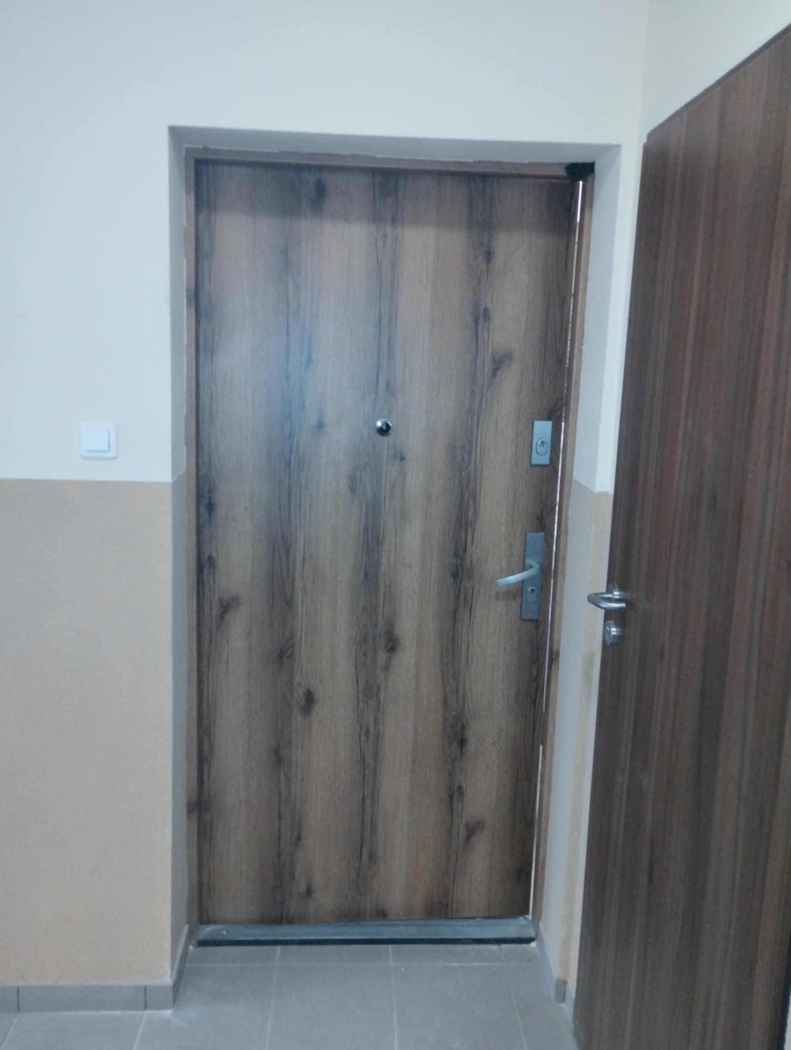 Zamknięte, brązowe drzwi wejściowe z wizjerem i srebrną klamką, imitujące drewno, w bloku mieszkalnym. Obok widoczne fragmenty drugiej pary drzwi.