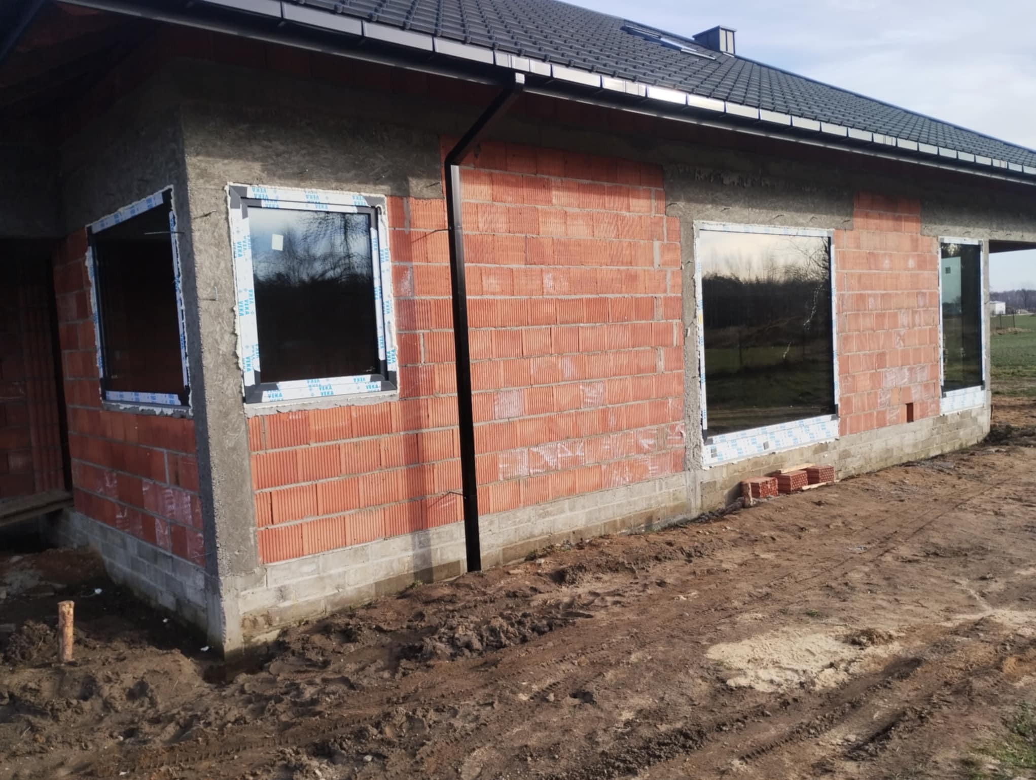 Surowy stan budynku z zamontowanymi oknami PCV marki Veka. Widoczne cegły i przygotowane otwory okienne. Teren wokół budynku jest ziemisty.