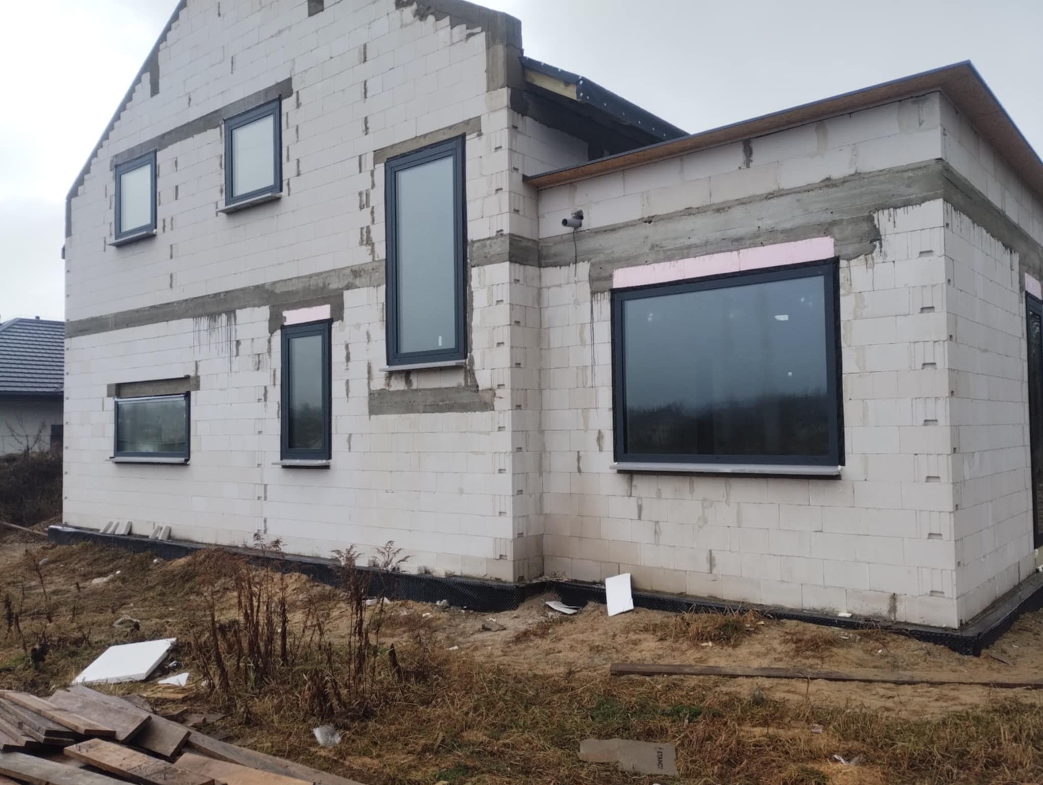 Surowy stan domu z zamontowanymi oknami w grafitowych ramach. Częściowo ocieplone otwory okienne. Widoczne bloczki z betonu komórkowego i teren budowy.