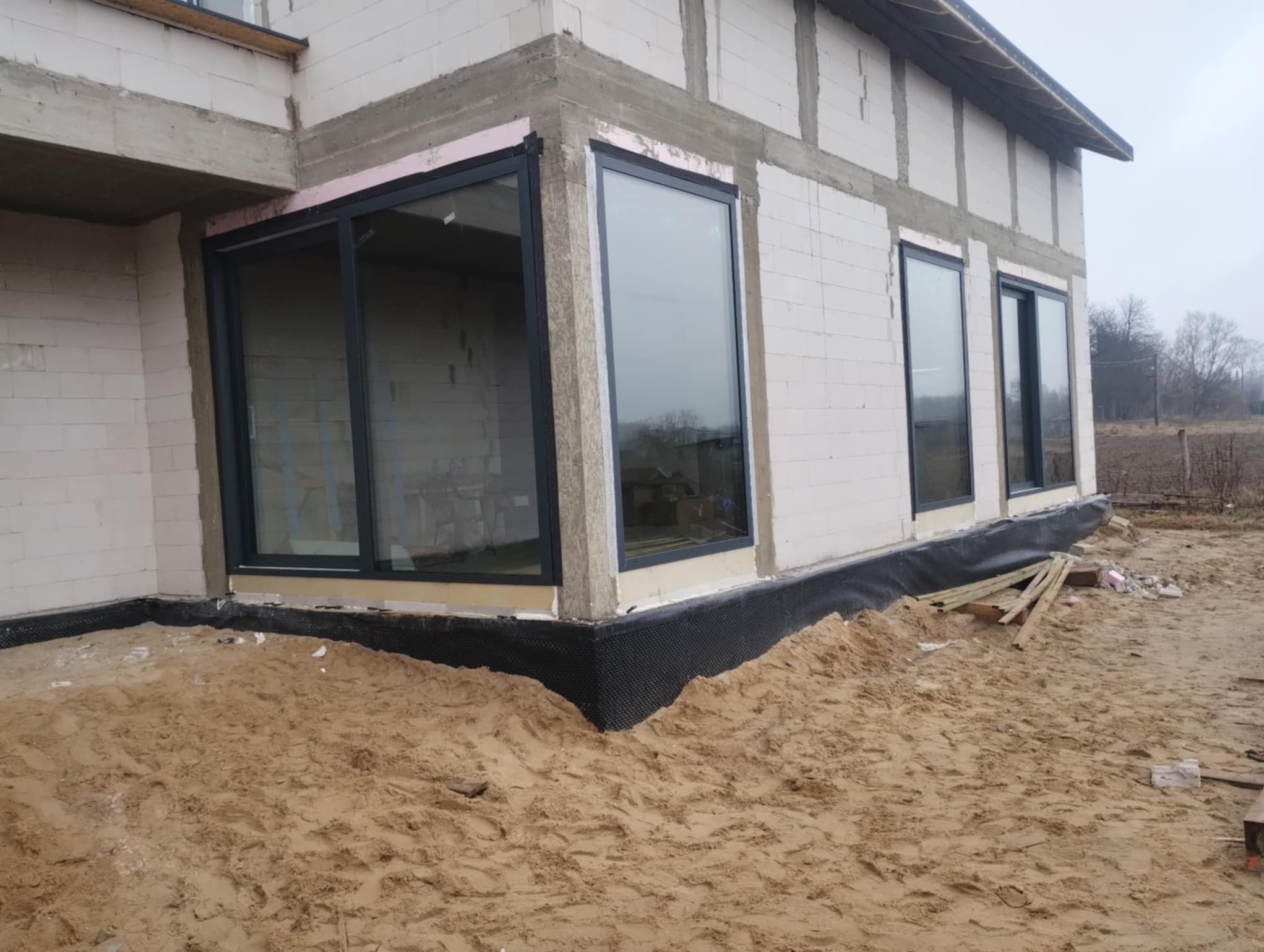 Surowy stan budynku z zamontowanymi oknami z czarnymi ramami. Widoczne ocieplenie i czarna folia fundamentowa. Na pierwszym planie piasek.