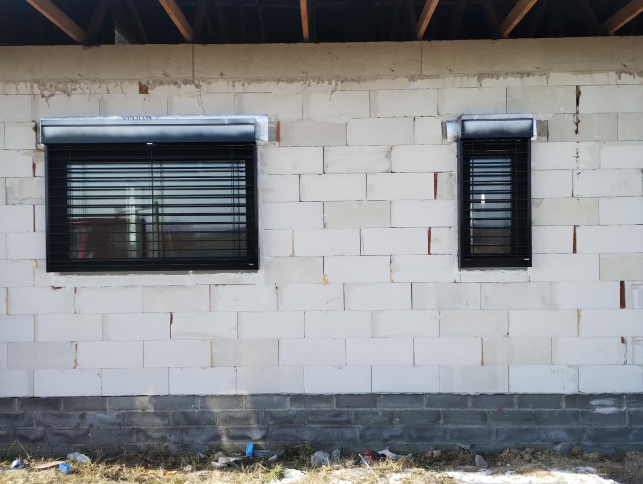 Dwa okna z roletami zewnętrznymi na surowej ścianie z betonu komórkowego. Rolety w kolorze czarnym, częściowo opuszczone. Widoczne niedokończone elementy budynku.