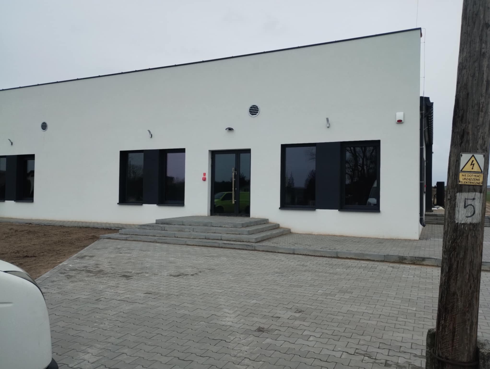 Biały budynek z czarnymi oknami i drzwiami wejściowymi, widok z zewnątrz. Kostka brukowa przed budynkiem. Słup energetyczny z ostrzeżeniem po prawej.