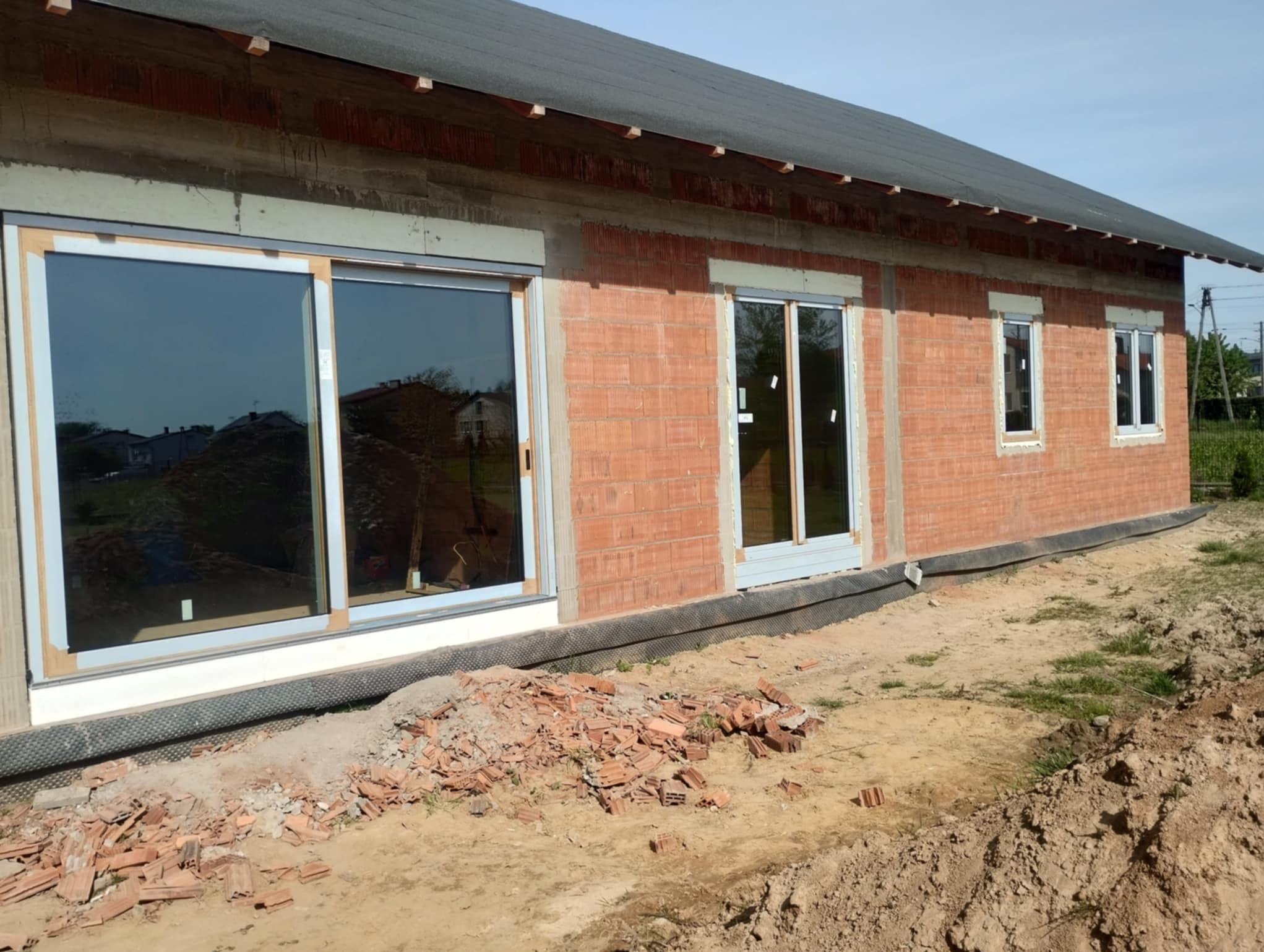 Surowy budynek z zamontowanymi oknami PCV i drzwiami balkonowymi. Widoczne cegły i elementy konstrukcyjne. Na pierwszym planie gruz i piasek.