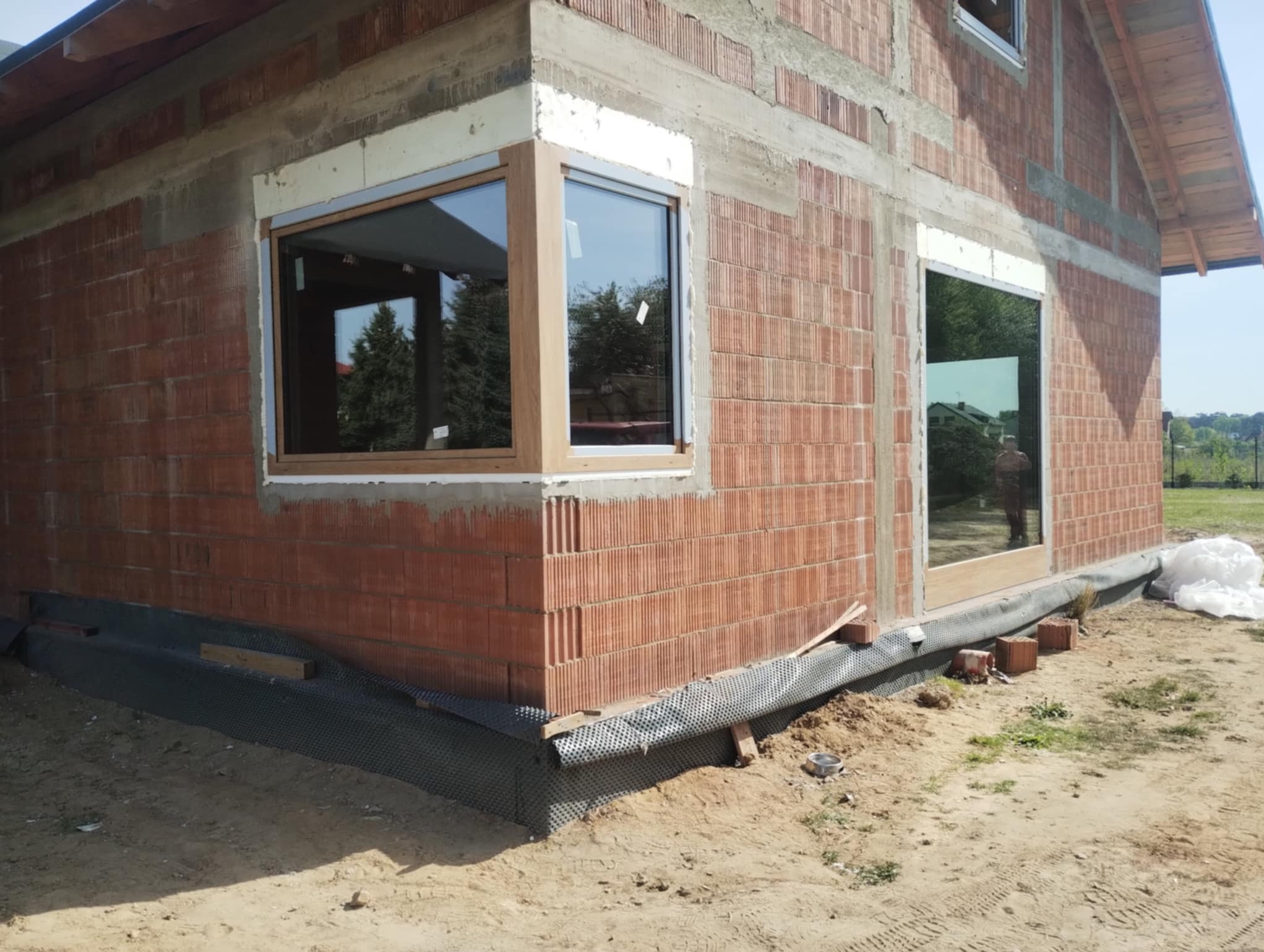 Surowy stan budynku z zamontowanymi oknami z drewnianymi ramami. Widoczne bloczki ceramiczne i warstwa izolacji fundamentów. W tle zieleń i domy.