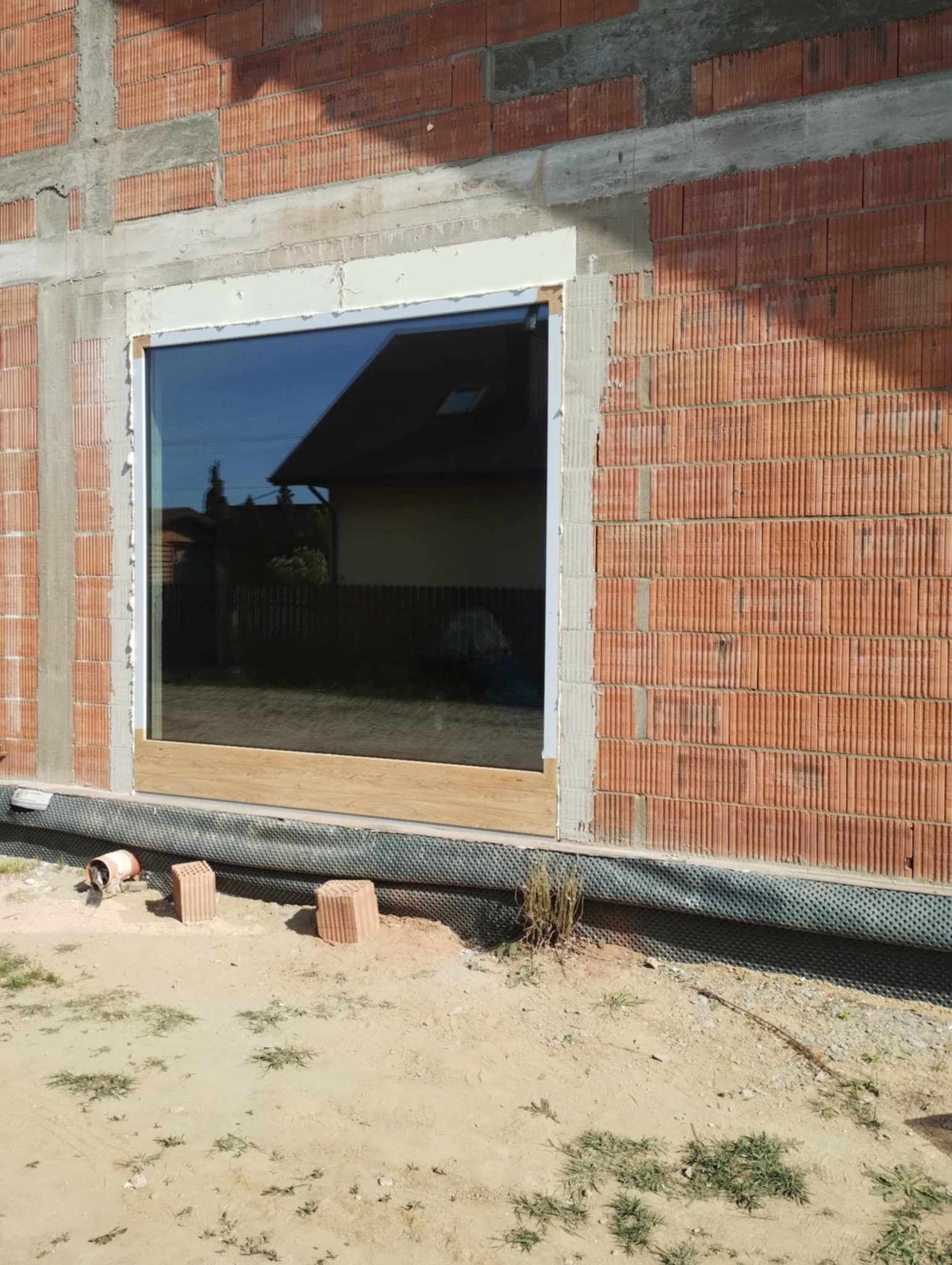 Wstawione okno w surowym murze z czerwonej cegły. Widoczna rama okienna z pianką montażową. W szybie odbija się budynek. Dół okna zabezpieczony deską.