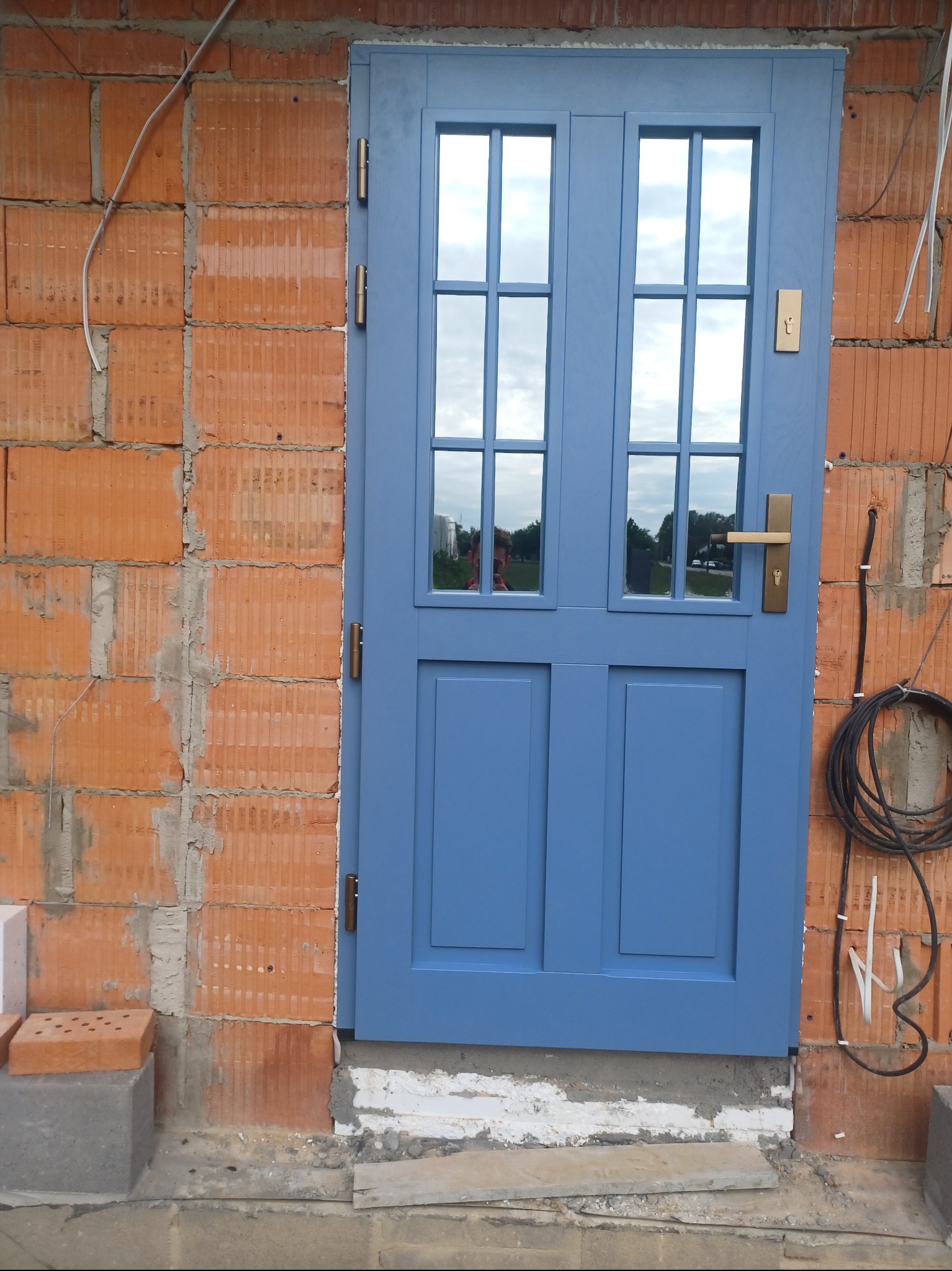 Niebieskie drzwi zewnętrzne z szybami, osadzone w surowym murze z cegły. Widoczne przewody instalacyjne i fragment otoczenia odbijający się w szybach.