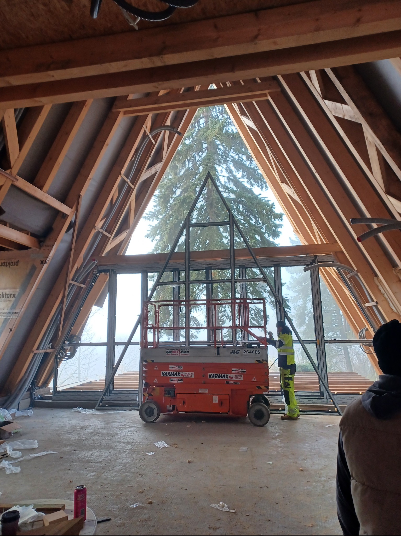 Montaż okien w nietypowym, trójkątnym otworze dachowym. Pracownik na podnośniku, widok na zewnątrz. Stalowa konstrukcja okien. Surowy stan wykończenia.