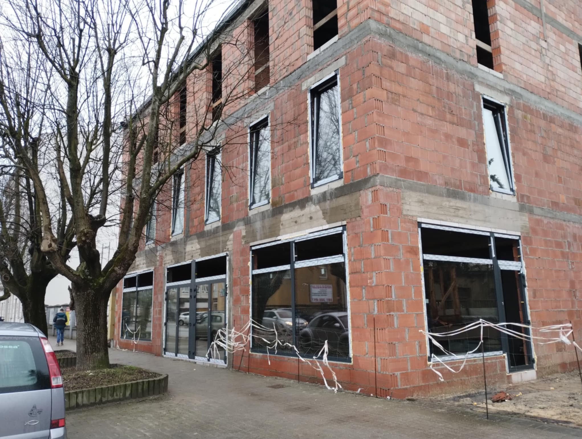 Budynek w trakcie montażu okien PCV. Widoczne surowe ściany z cegły, częściowo zamontowane okna z czarnymi ramami, drzewa i fragment chodnika. Etap prac budowlanych.