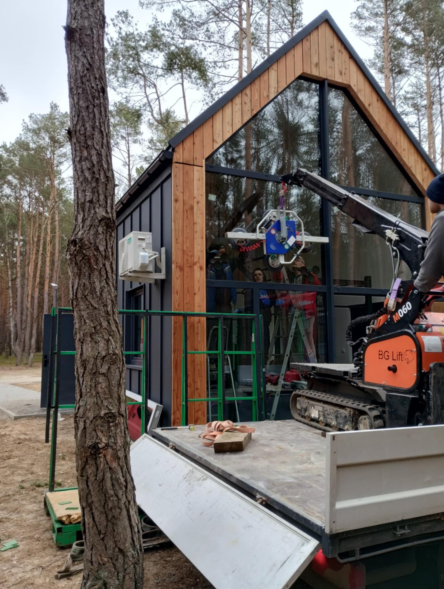 Montaż okna fasadowego w budynku o konstrukcji szkieletowej. Na pace samochodu dźwig do szyb, a w tle las.