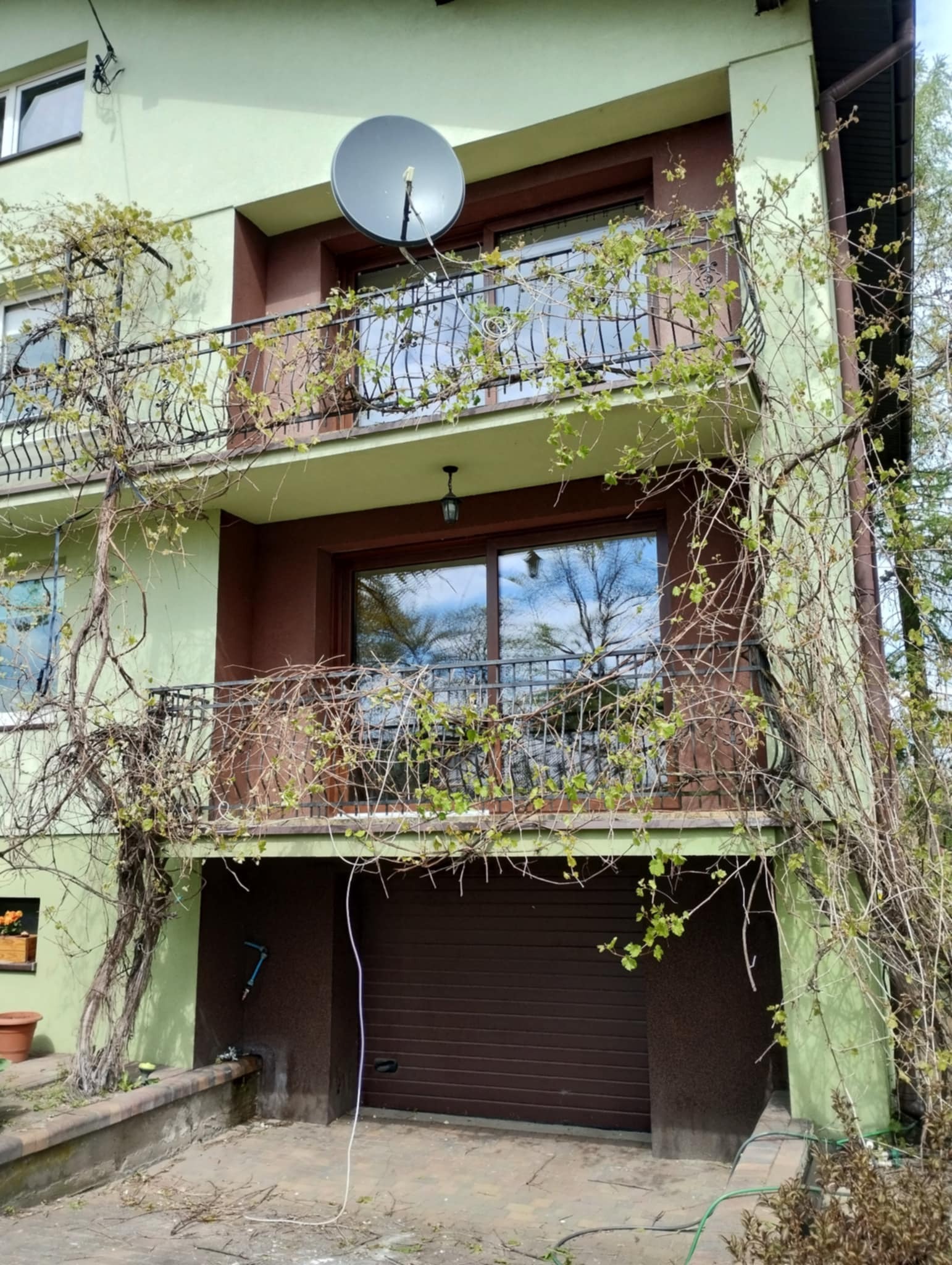 Dwupiętrowy dom z balkonami i garażem. Na balkonach ozdobne, metalowe balustrady oplecione pnączami. Na górnym balkonie antena satelitarna. Garaż z brązową bramą.
