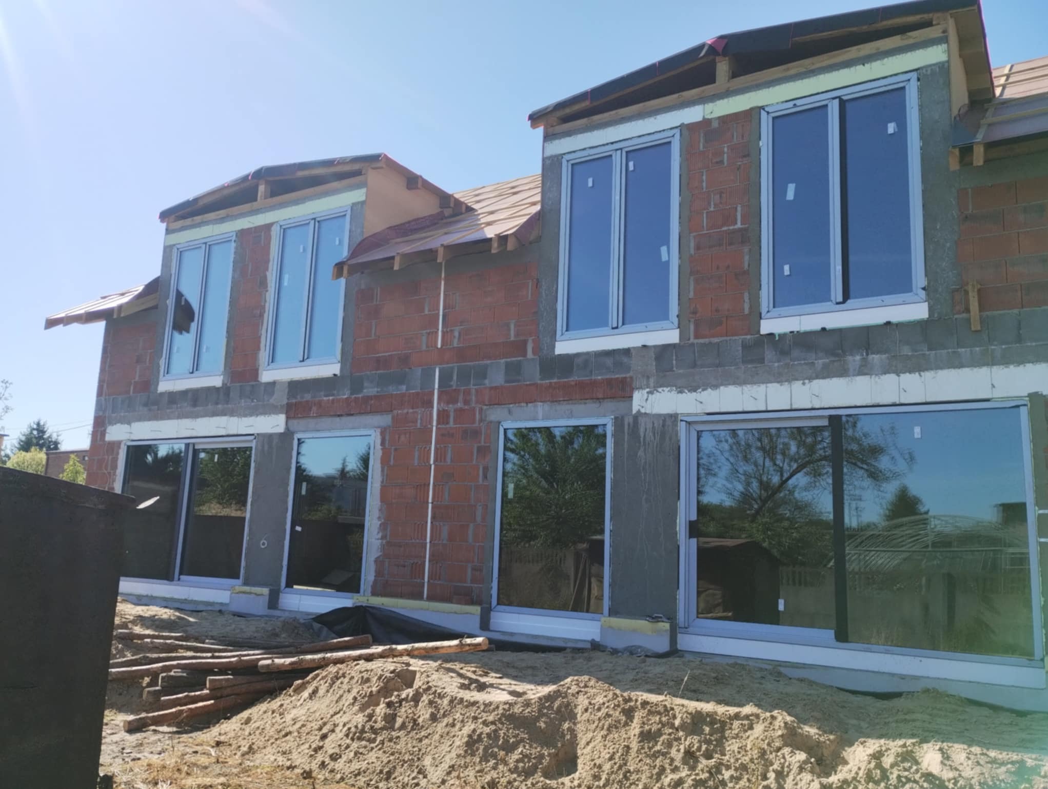 Wznoszony budynek z zamontowanymi oknami PCV. Widoczne ślady prac murarskich i przygotowania do dalszych etapów budowy. Okna z wyraźnymi refleksami nieba i drzew.