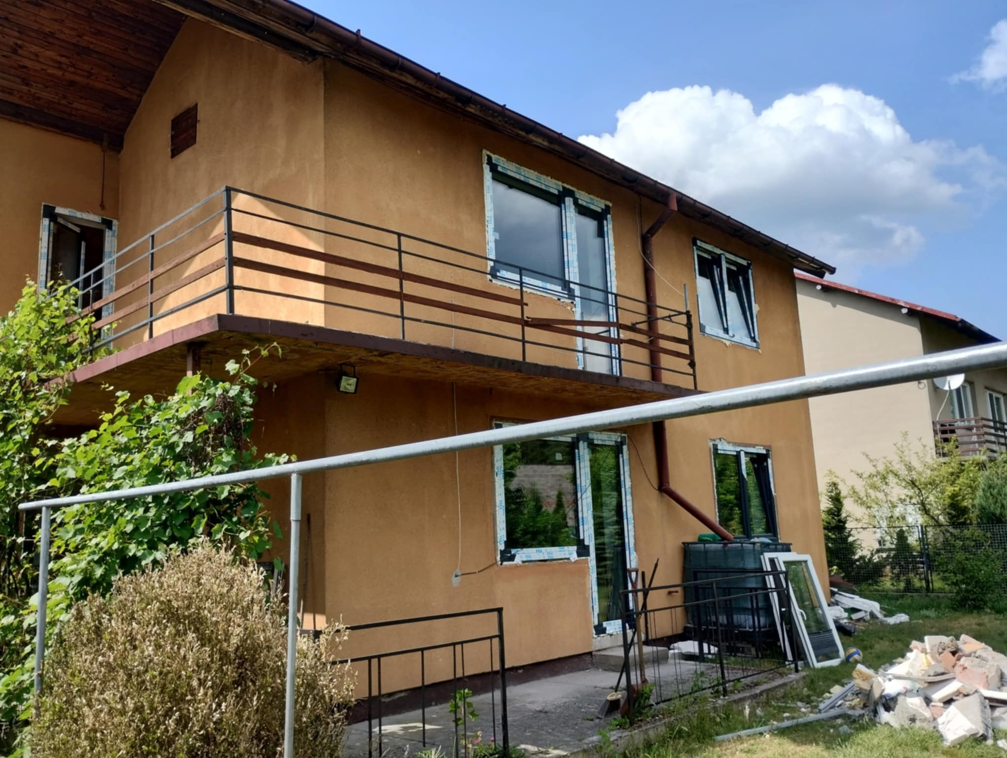 Dom z balkonem i oknami w trakcie montażu. Widoczne narzędzia i odpady budowlane. Częściowo zamontowane okna z folią ochronną. Słoneczny dzień.