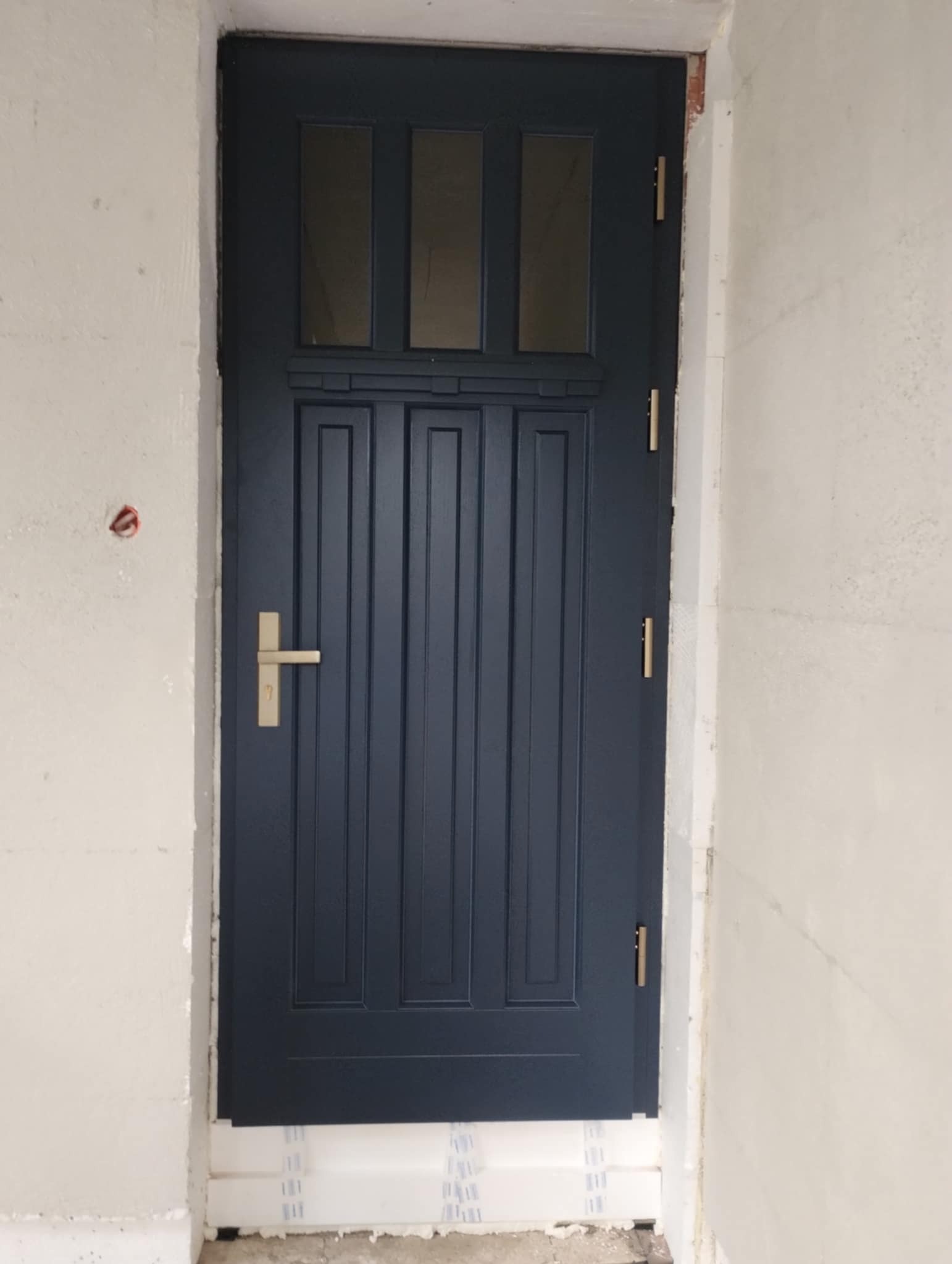 Granatowe drzwi z trzema szybkami w górnej części, złota klamka i zawiasy, osadzone w surowym otworze ściennym z widoczną izolacją.