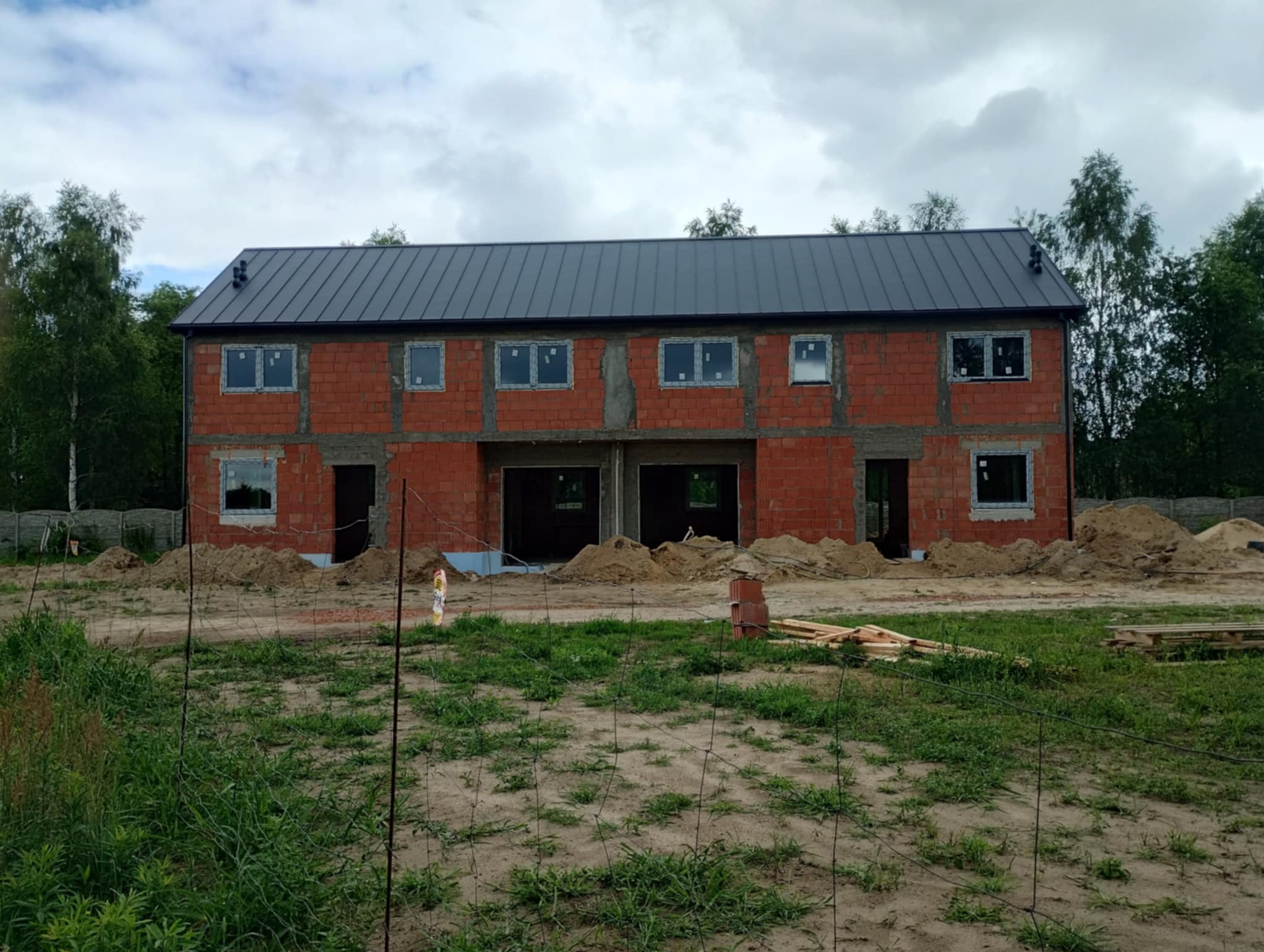 Surowy stan bliźniaka z oknami w Zduńskiej Woli, ceglana elewacja, szare okna i dach, teren przygotowany pod budowę, widok z zewnątrz na tle zieleni.