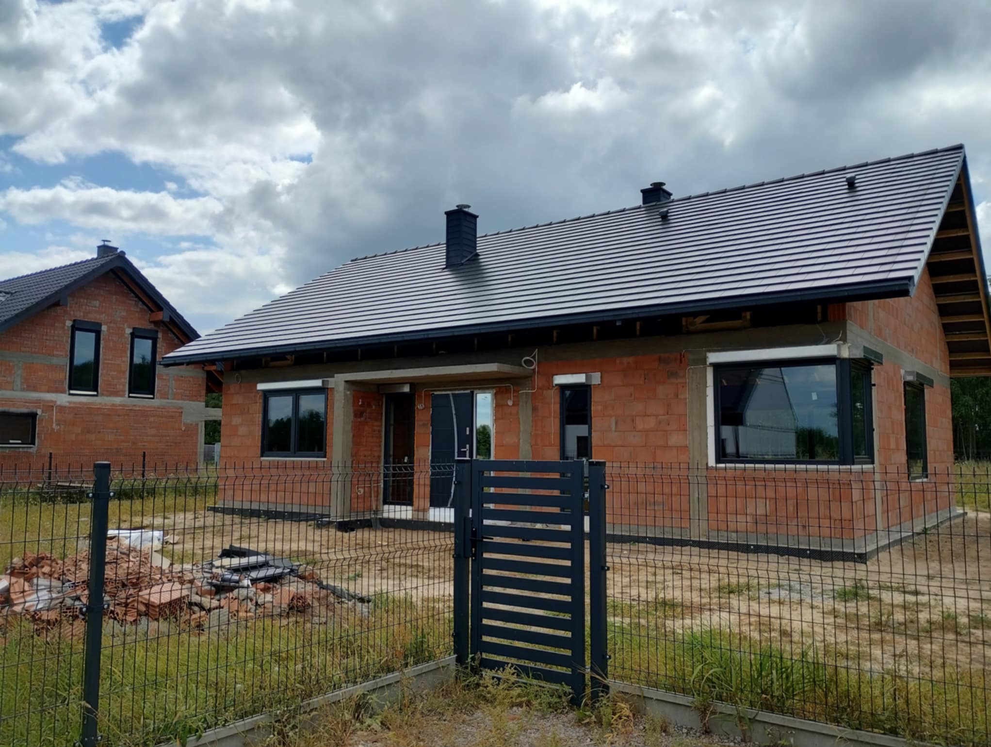 Surowy dom z czarną stolarką okienną i roletami zewnętrznymi, ogrodzony siatką. Widoczne fragmenty dachu i kominy. W tle drugi budynek w stanie surowym.