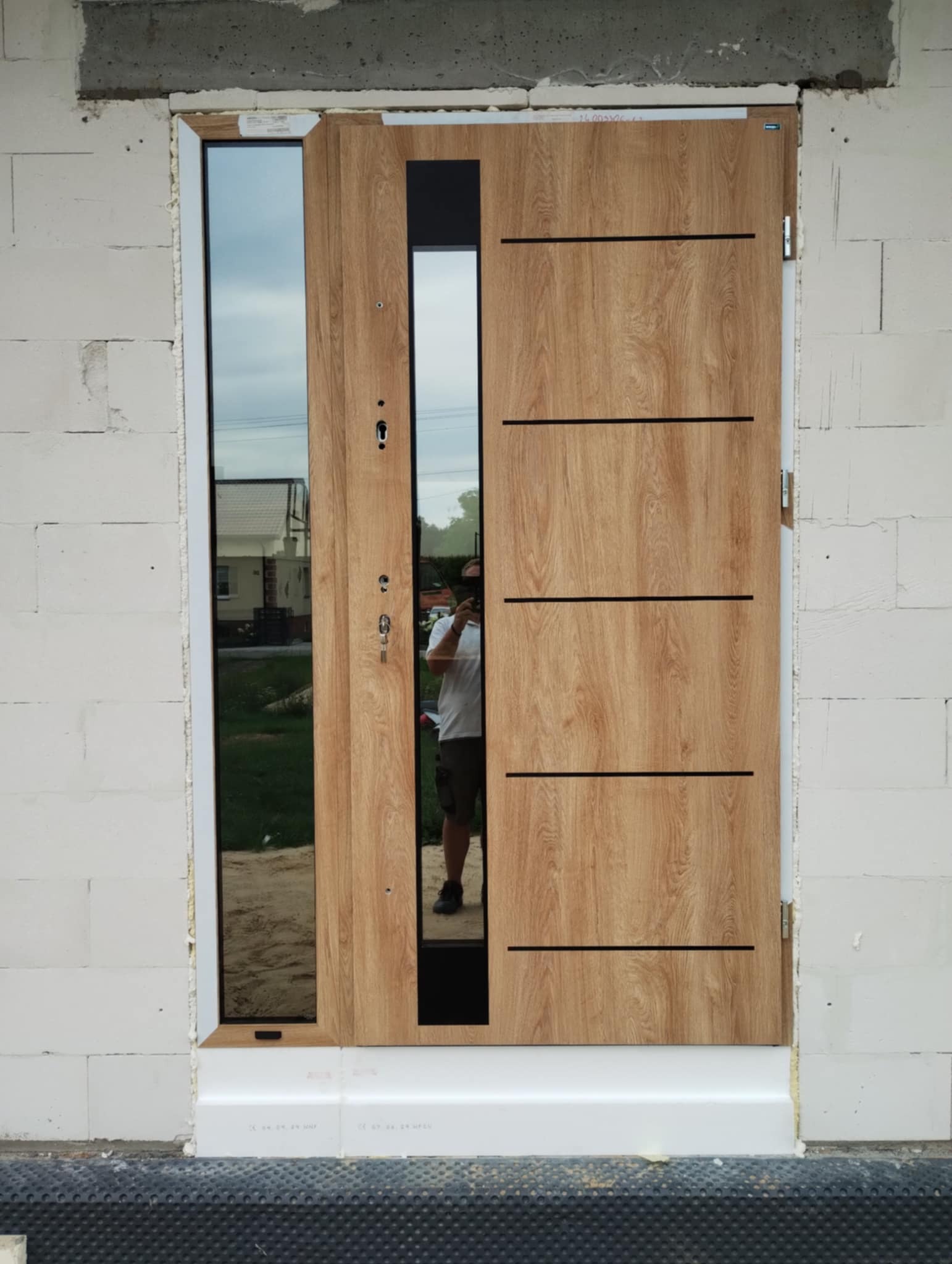 Drewnopodobne drzwi wejściowe z szybą, zamontowane w surowym murze. Widoczne detale: zamek, klamka, poziome frezy. W szybie odbija się osoba robiąca zdjęcie.