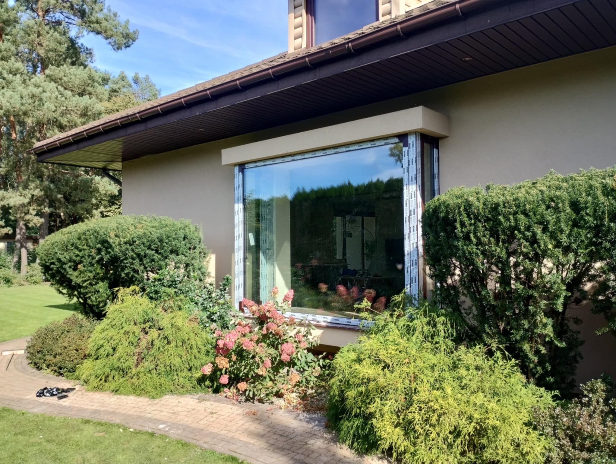 Zamontowane okno PCV w elewacji domu z brązowym dachem, otoczone zielenią krzewów i drzew, widoczne z zewnątrz na tle błękitnego nieba.