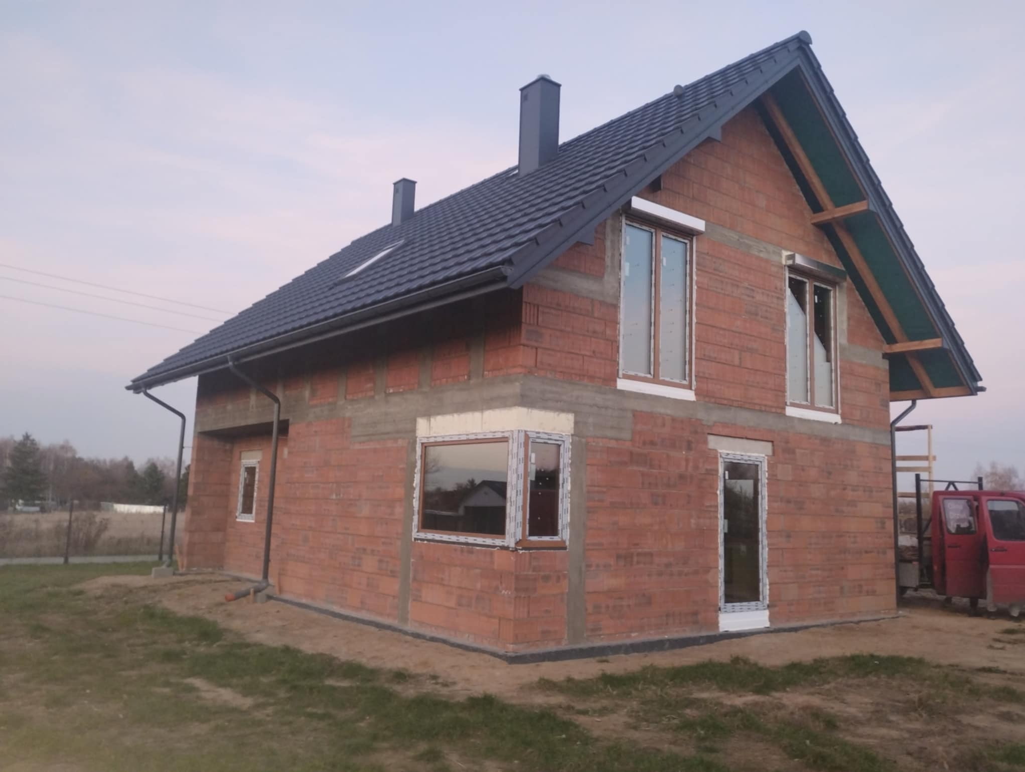 Surowy stan domu jednorodzinnego z czerwonej cegły z zamontowanymi oknami PCV i drzwiami. Widoczne elementy dachu i rynny. Na zewnątrz zaparkowany czerwony samochód dostawczy.