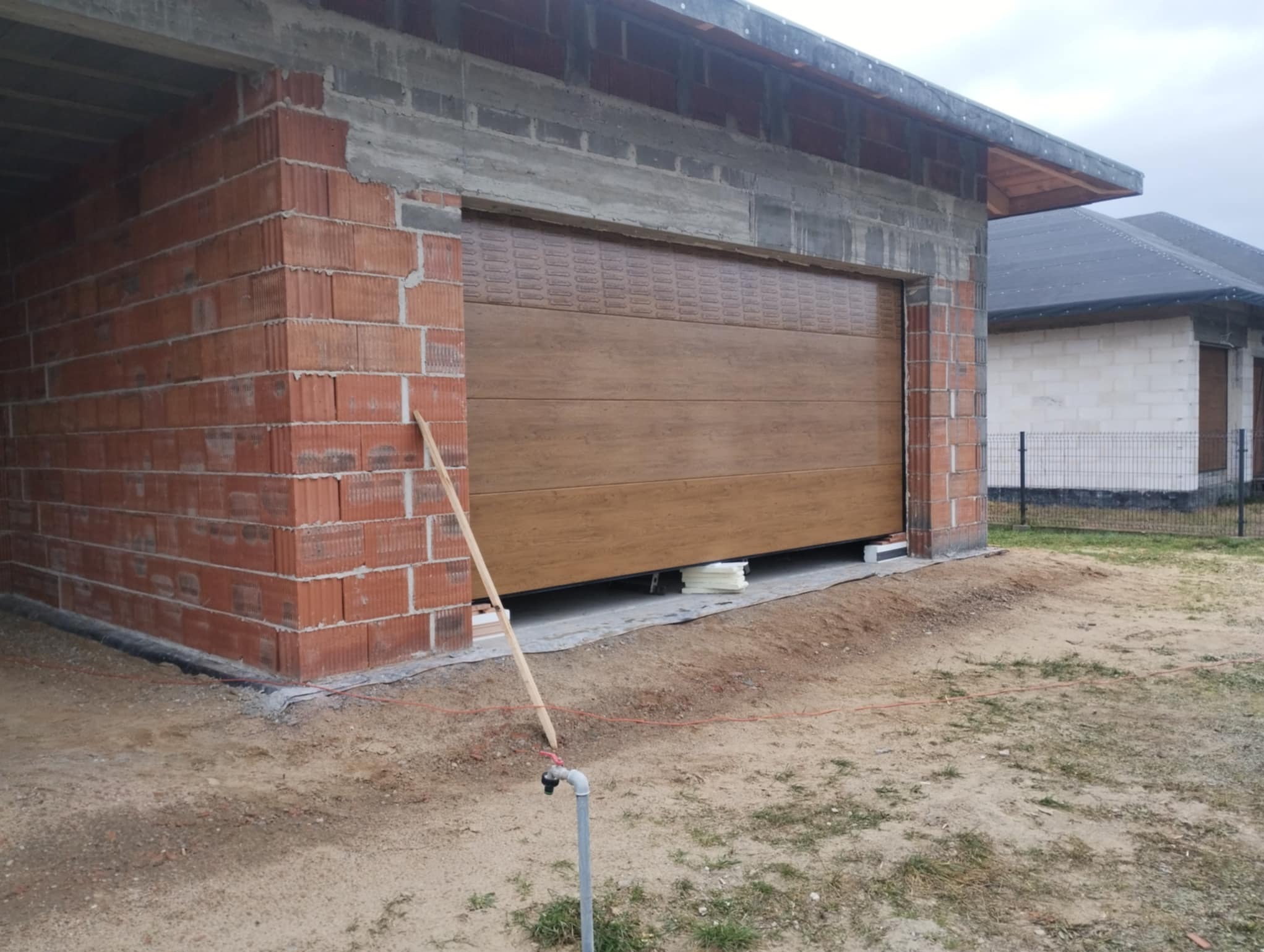 Montaż brązowej bramy garażowej segmentowej w nowym budynku z czerwonej cegły. Brama częściowo otwarta, widoczne elementy montażowe i materiały izolacyjne.