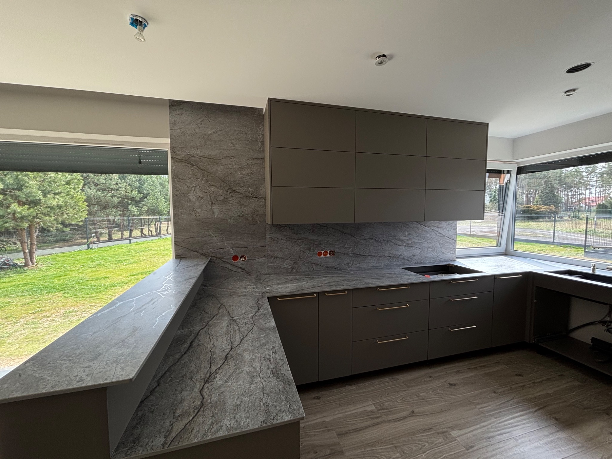 Nowoczesna kuchnia z szarymi szafkami i blatem z kamienia naturalnego o wyraźnym użyleniu. Duże okno z widokiem na zieleń. Minimalistyczny design.