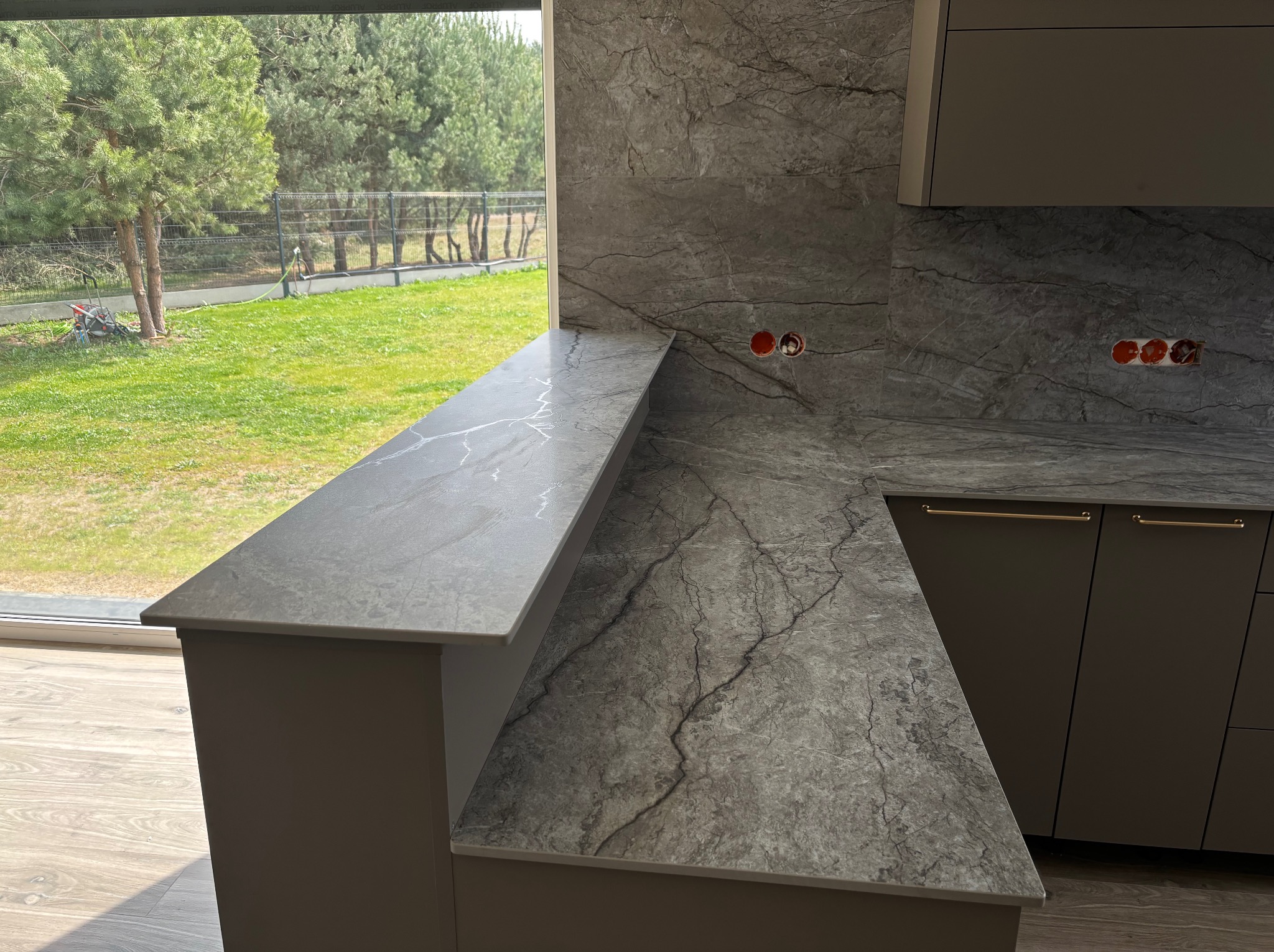 Szary blat kamienny z wyraźnym wzorem żyłkowania, zamontowany w kuchni z widokiem na zieleń za oknem. Widoczne gniazdka elektryczne na ścianie.