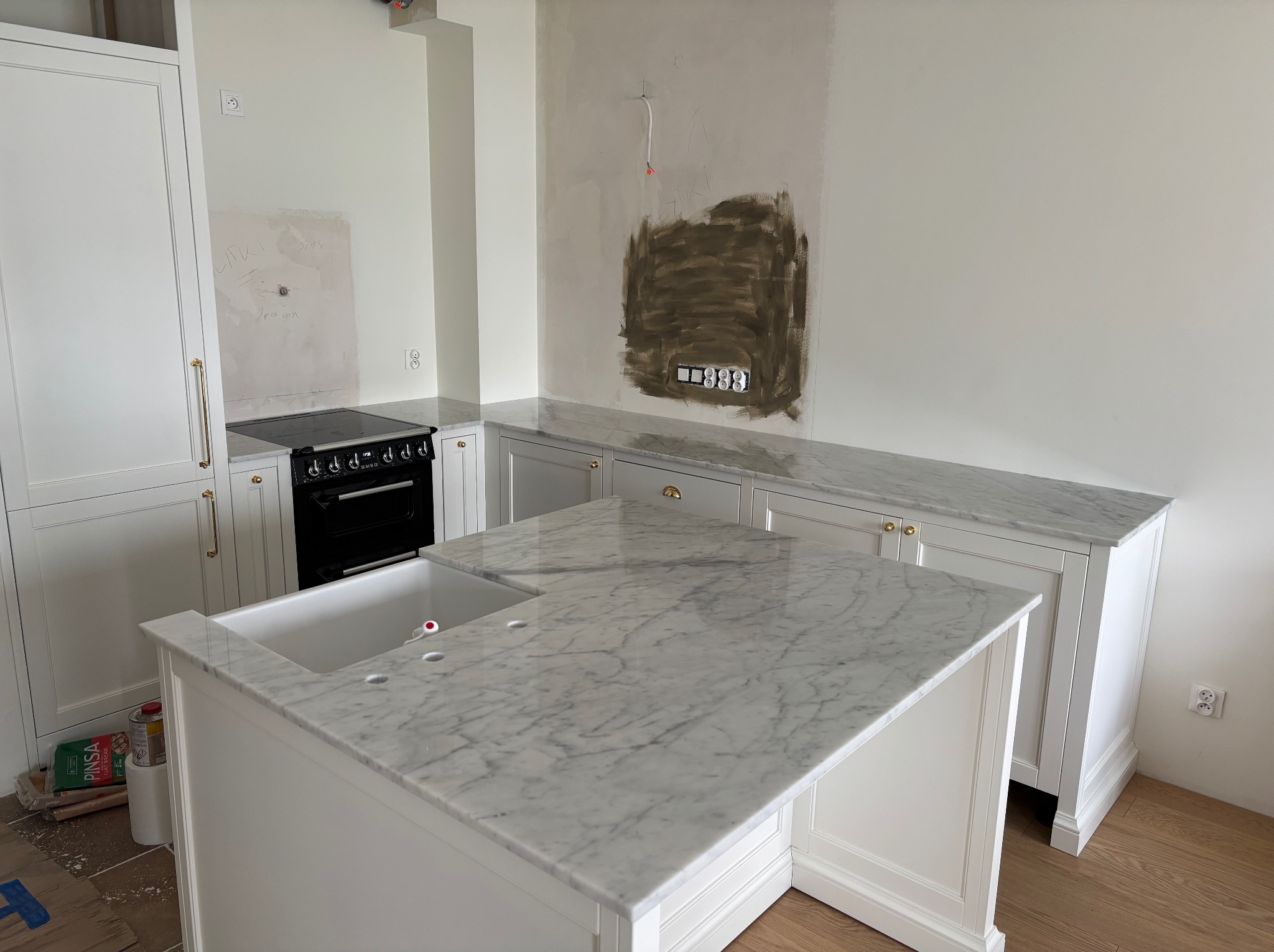 Biała wyspa kuchenna z marmurowym blatem w klasycznej kuchni. Widoczne przygotowania do montażu osprzętu elektrycznego na ścianie.