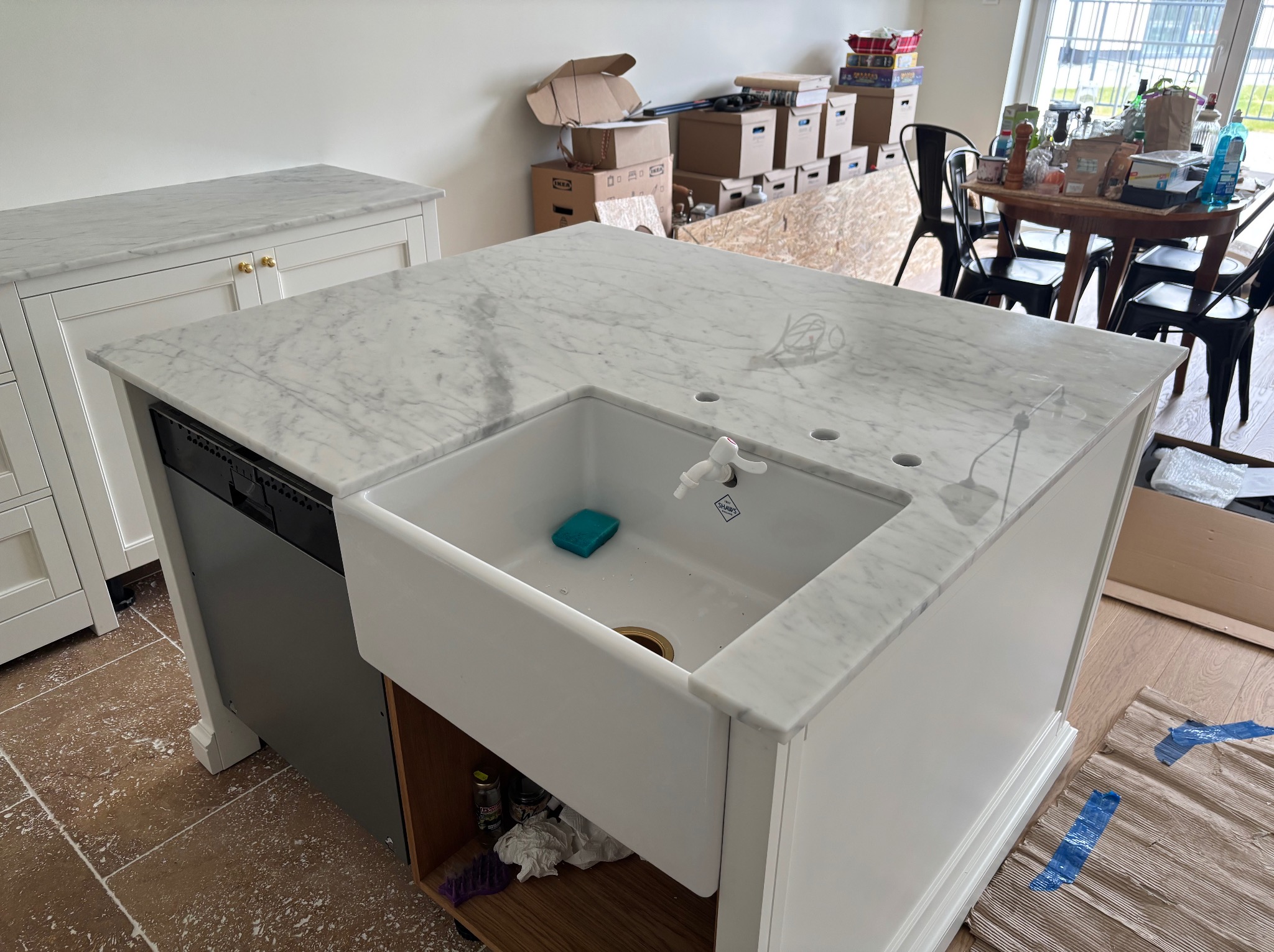 Wyspa kuchenna z blatem z białego marmuru i ceramicznym zlewem farmerskim. Widoczne otwory na baterię i dozownik. W tle kartony i stół z krzesłami.